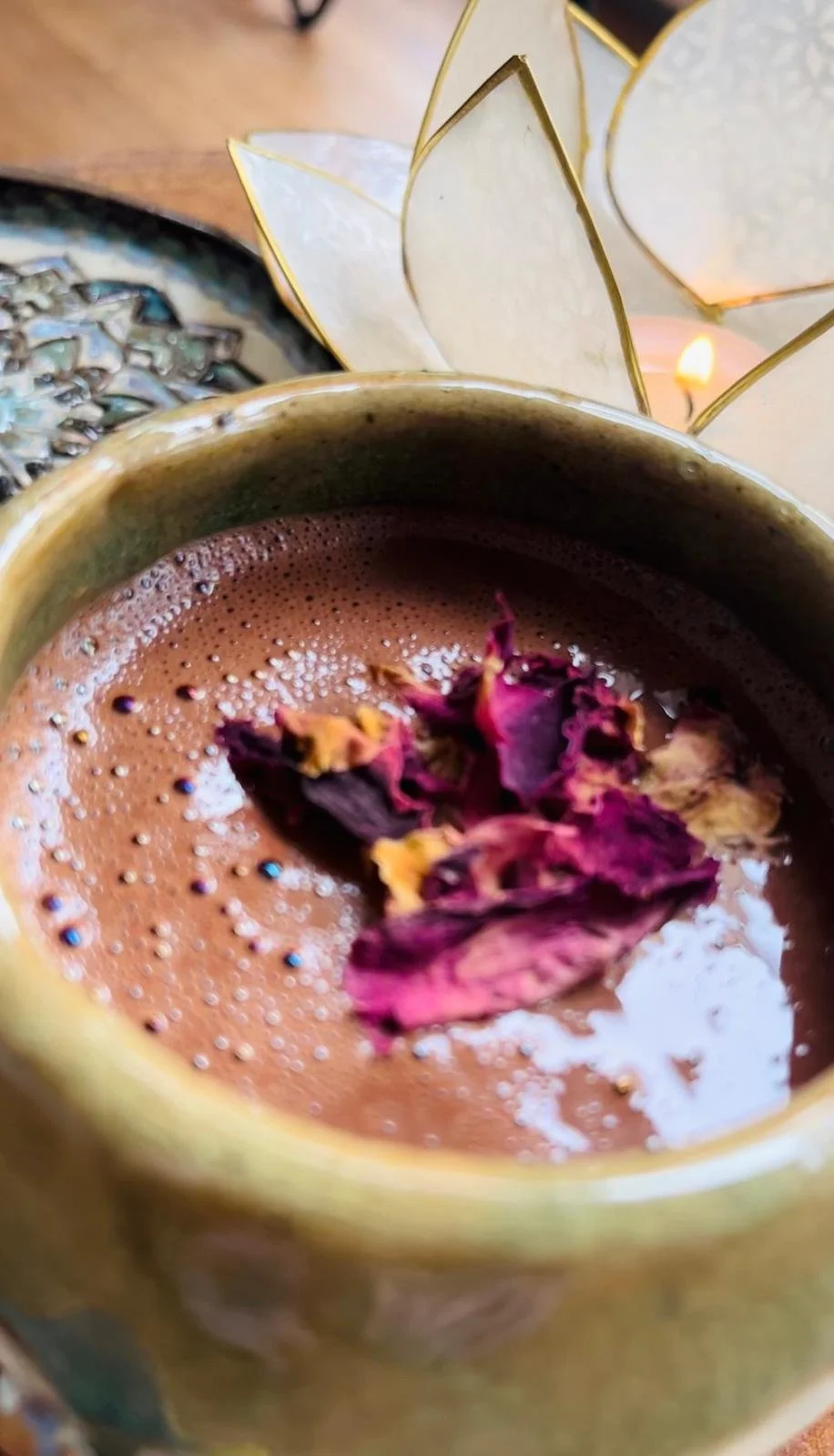 Eine Tasse heiße Schokolade mit Trockenblumen als Dekoration, im Hintergrund eine Kerze und romantische Dekorationen.