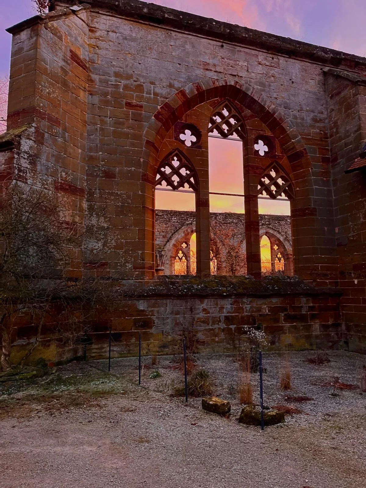 Rückansicht einer alten Kirche mit großen, spitzbogigen, gotischen Fenstern aus rotem Stein, bei Sonnenuntergang, mit verblasstem Himmel und Bäumen im Hintergrund.