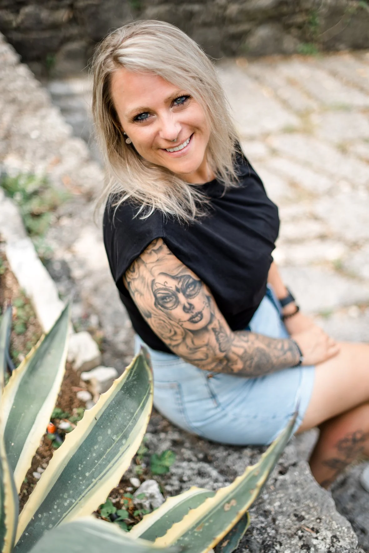 Junge Frau mit blonde, schulterlange Haare, tätowierter Arm und Piercings, sitzt in einer Gärtnerei oder einem Garten mit großen Agavenpflanzen, lächelt und schaut in die Kamera.