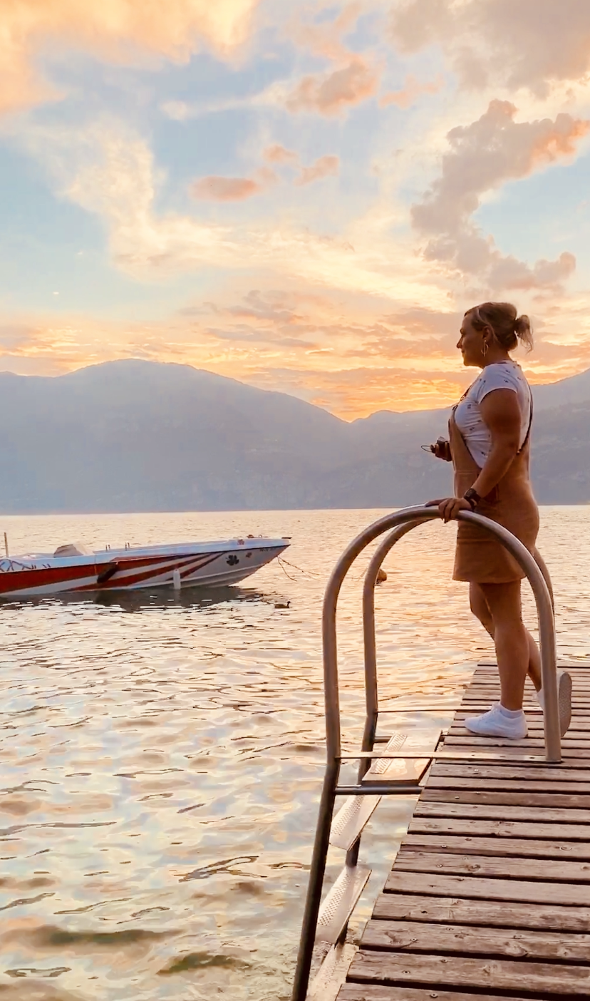 Eine Frau steht auf einem Steg am See und betrachtet ein Boot, während die Sonne untergeht und der Himmel in warmen Farben erstrahlt.
