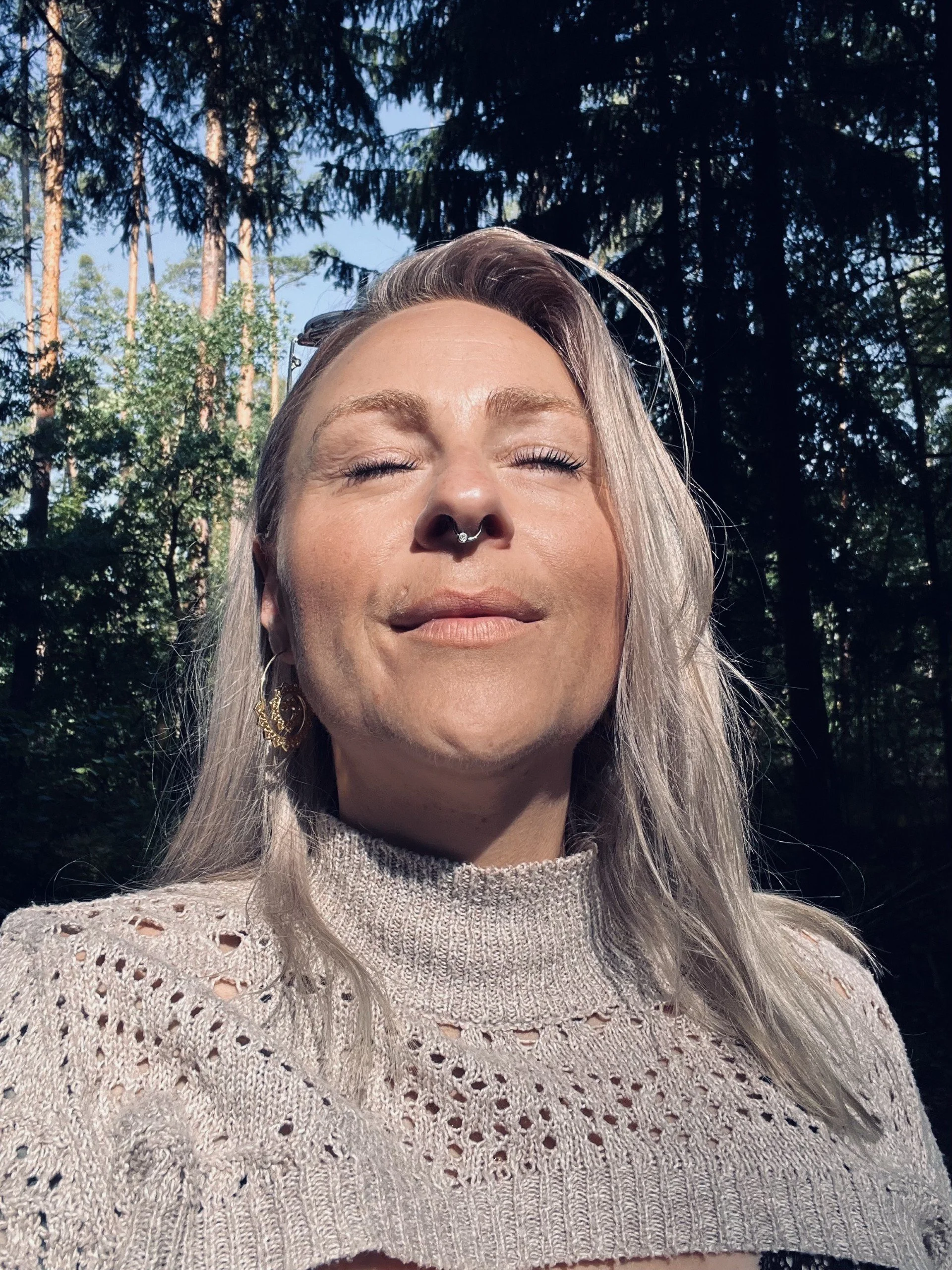 Junge Frau mit langen, grauen Haaren, geschlossenen Augen, Piercing an der Nase, Blume im Ohr, trägt eine Strickjacke und Sonnenbrille auf dem Kopf, im Wald bei daylight.