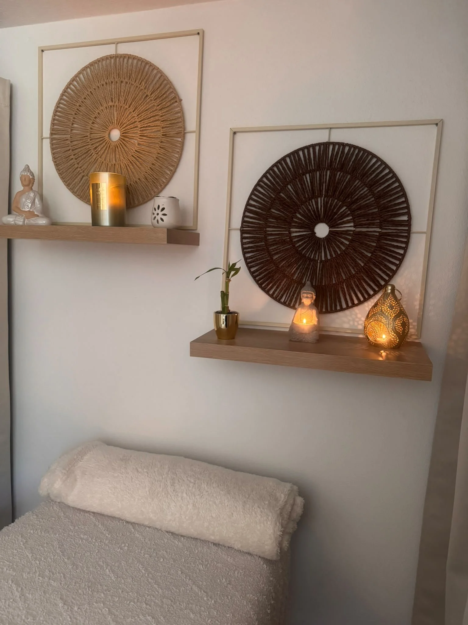 Ein Schlafzimmer mit weißen Wänden und zwei schwebenden Holzregalen. Auf dem linken Regal befindet sich eine kleine Buddha-Statue, ein Kerze und eine Keramikvase. Das rechte Regal beherbergt eine kleine Pflanze, eine beleuchtete Buddha-Figur und eine äthiopische Laterne. Unter den Regalen liegt ein großes, braunes, flauschiges Kissen auf einem Bett.