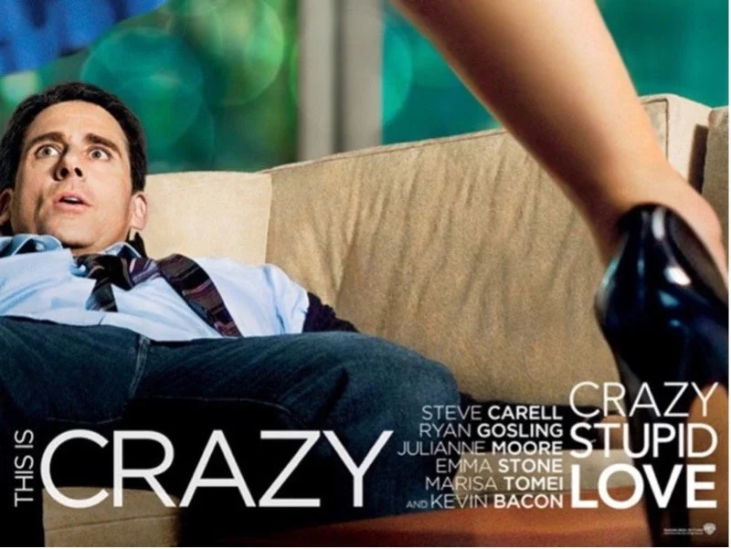 A man with dark hair, wearing a light blue shirt and a dark tie, is lying on a beige couch looking up with a surprised or shocked expression. A person's arm is reaching into the frame, holding a black shoe. Text on the image says "This is crazy, stupid love," with credits for the film.
