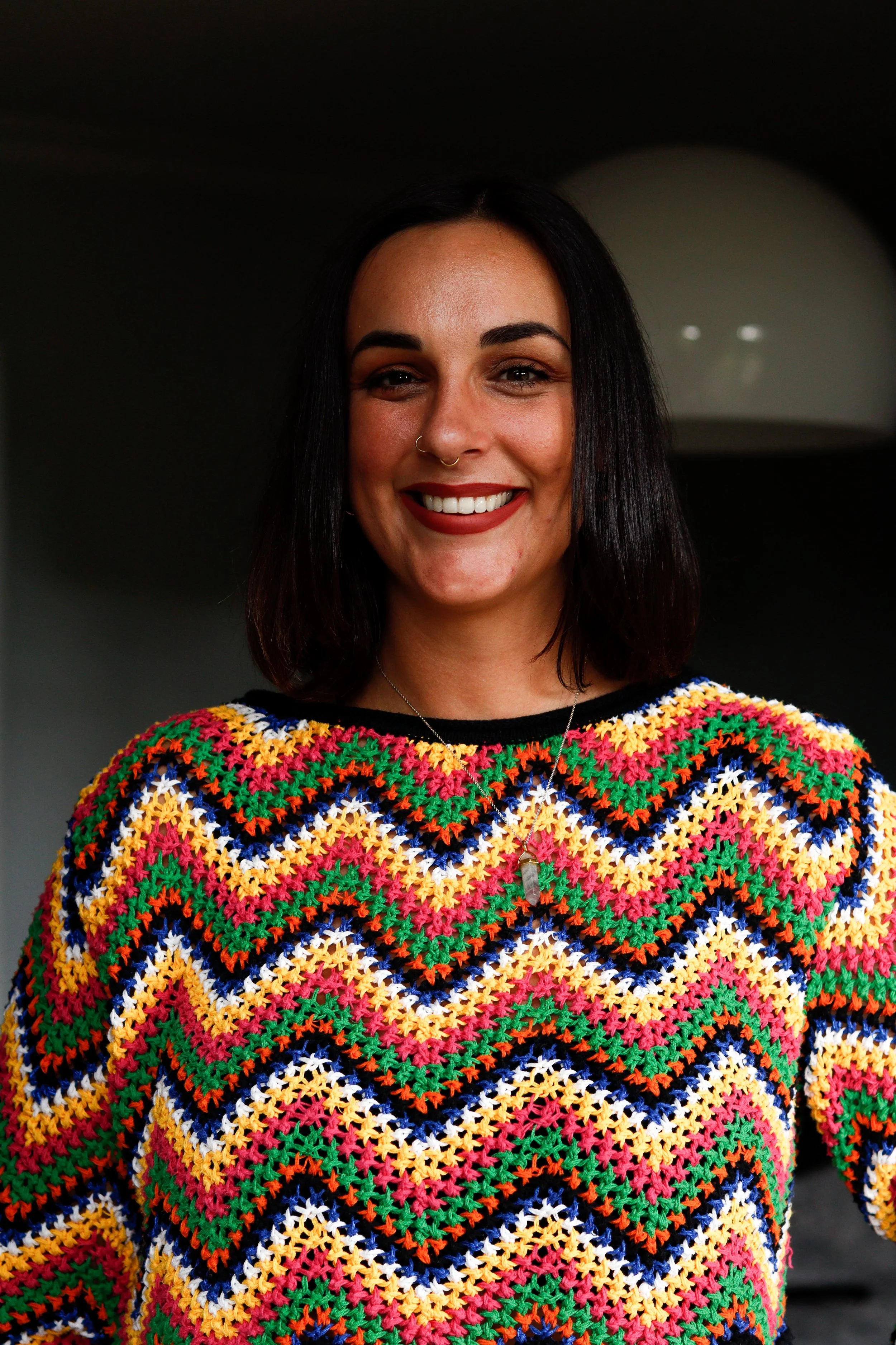 Close-up of a woman with dark hair and a nose ring, wearing a colorful crocheted sweater with a diamond pattern of various colors, smiling.