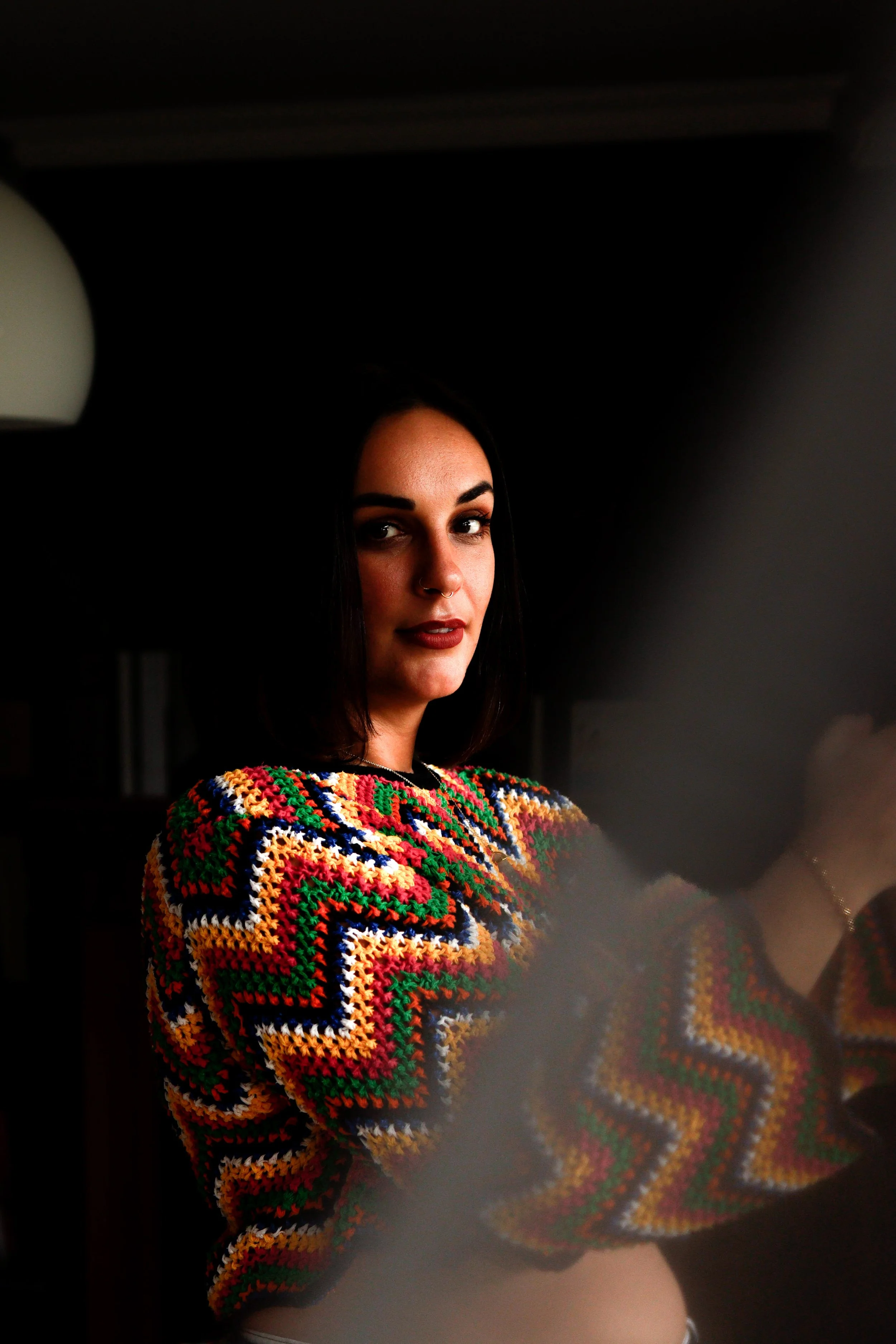 A woman with dark hair, dark eyebrows, and a septum piercing, wearing a colorful crocheted sweater, is looking slightly to the side against a dark background.