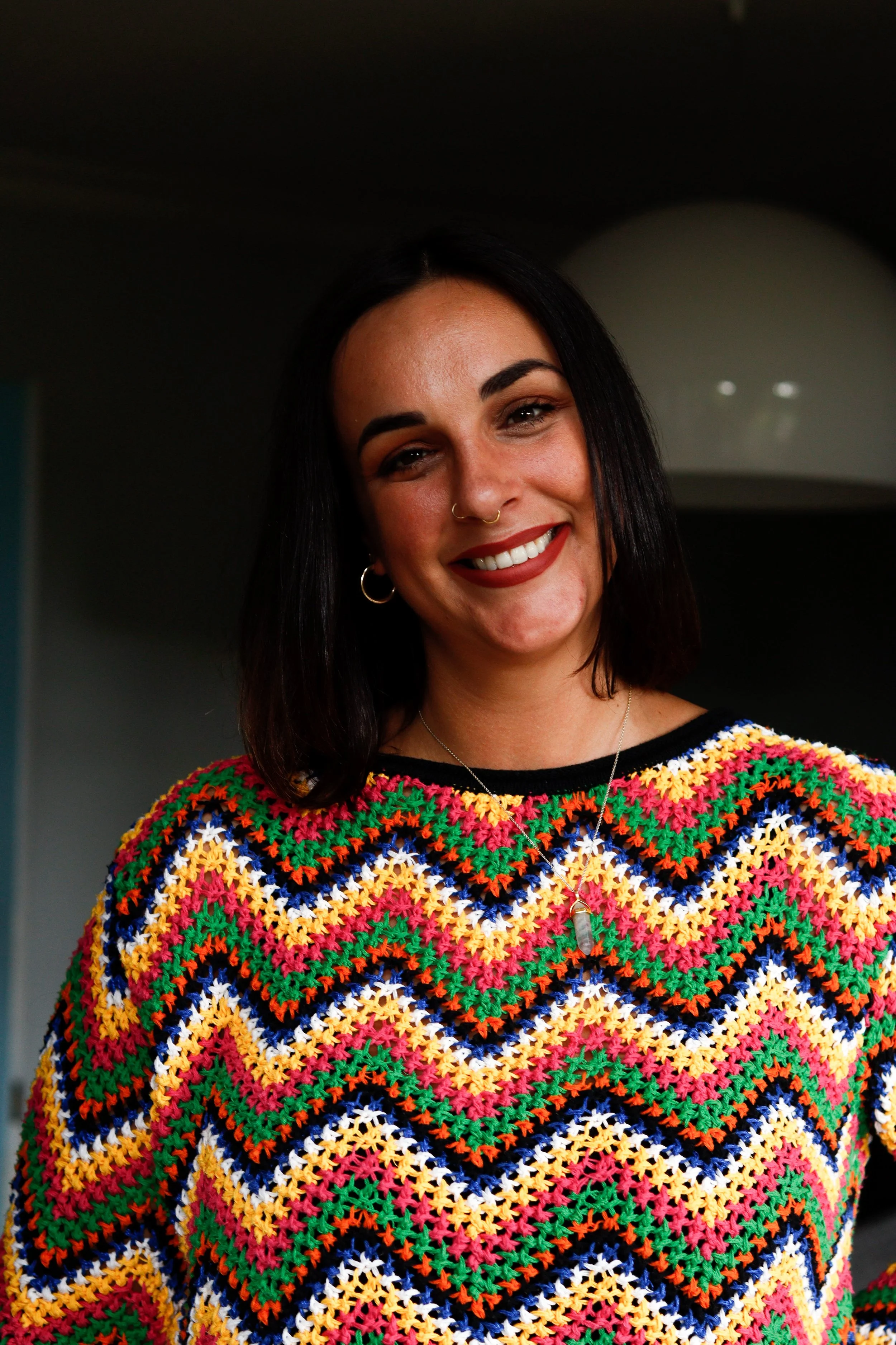 A woman with black hair and a nose ring wearing a colorful crochet sweater, smiling indoors.