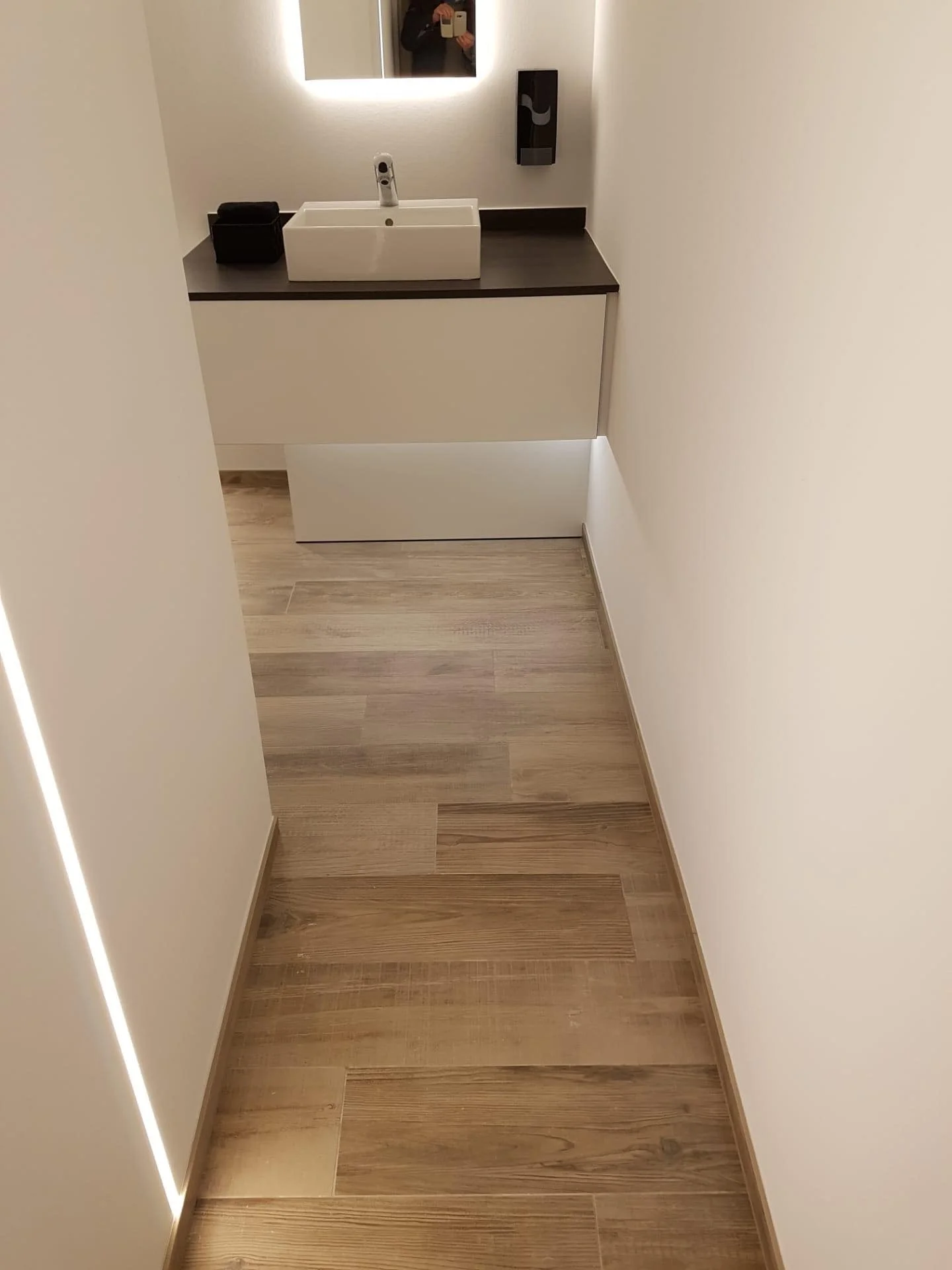 Klein, modern washokje met een witte wasbak, een spiegel met omgevingslicht, een zwart handdoekje of doek en een dispenser aan de muur, met houten vloer en witte muren.