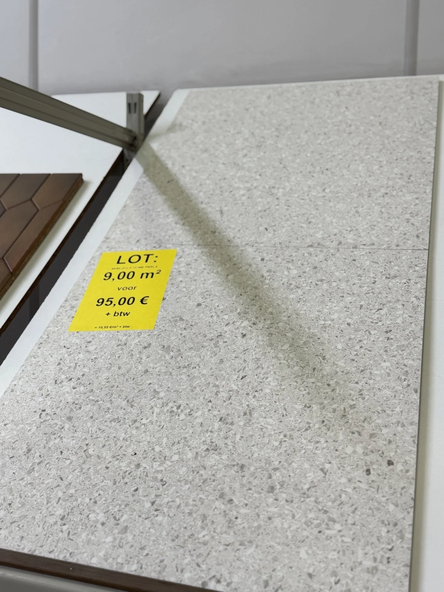 Een dichtgelegde vloer met een gele prijskaart die 9,00 m2 aangeeft voor 95,00 euro plus btw, met een stuk vloer en een stuk vinyl vloerbedekking ernaast.