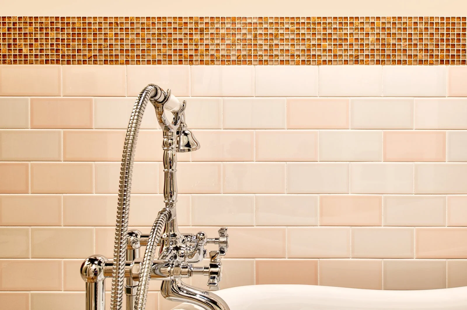 Badkamer met een wit bad, een chroom kraan en een hoofddouche, met wandtegels in lichtroze en een strook kleine bruine mosaïektegels boven de lichtroze tegels.