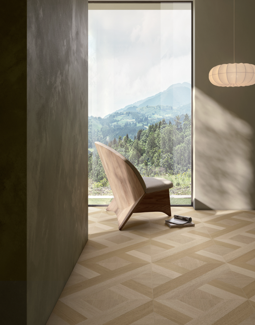 Een interior met een grote raam die uitzicht biedt op groene heuvels en bergen, een moderne houten stoel, een wandlamp, en enkele boeken en een tablet op de vloer.