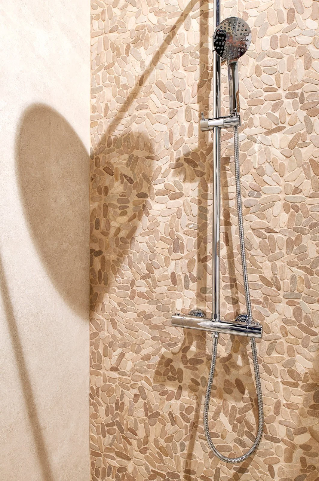 Douche met een ronde, chromen douchekop en een chroomhouder, geïnstalleerd op een muur met een decoratief tegelpatroon van kleine, ovale tegels in aardetinten.