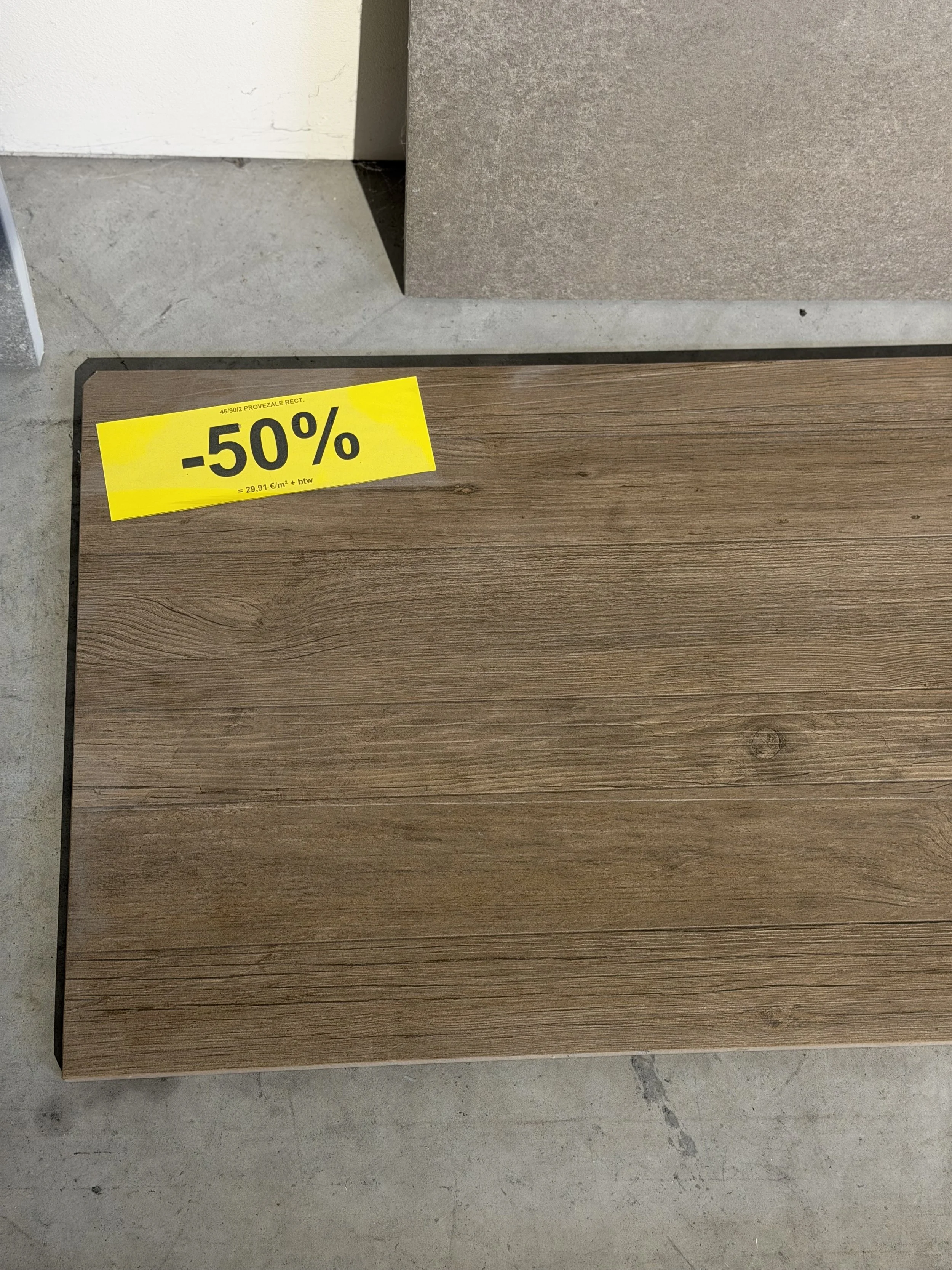 Een houten vloerplaat met een geel uitverkoopsticker die 50% korting aangeeft, boven een betonnen vloer en een muur bekleed met steen.