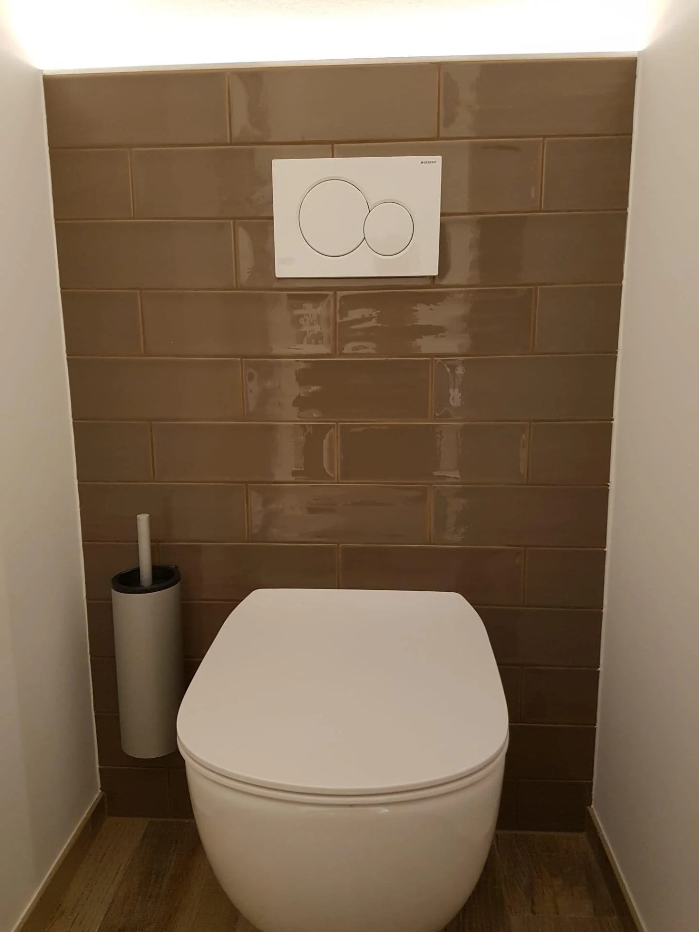 Wc met een witte toiletpot, een toiletrolhouder en een toiletborstel, met een bruin betegelde muur op de achtergrond.