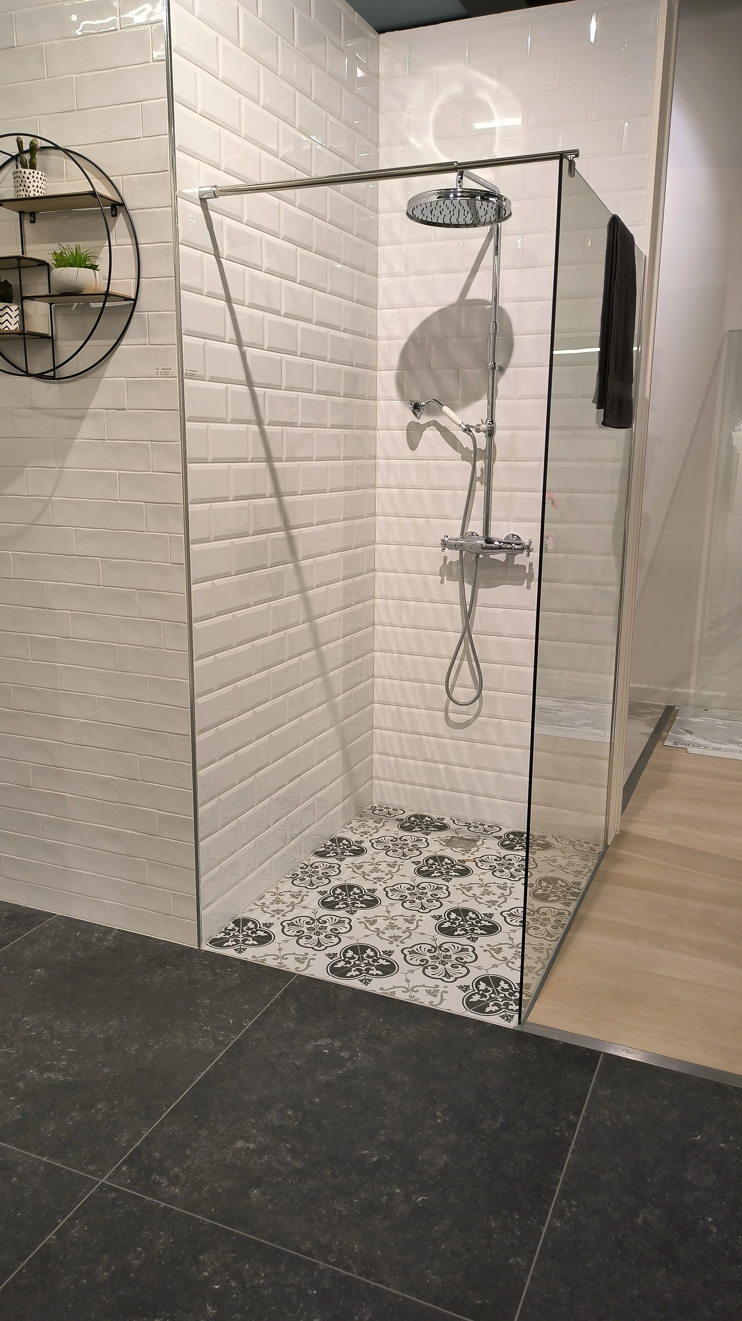 Inloopdouche met witte tegelwanden en decoratieve vloer tegels met zwarte bloemmotieven, inclusief regen douchekop en zwarte handdouche, gescheiden door een glazen wand.