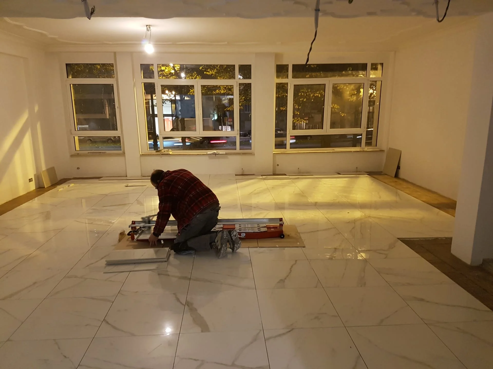Man in een nieuw ingerichte ruimte met grote ramen die uitkijken op straat, bezig met installatie of renovatie, met gereedschap en materialen op de vloer.