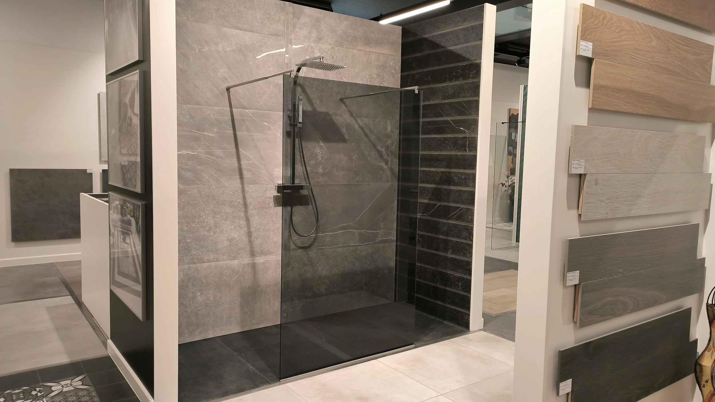 Een badkamer showroom met een inloopdouche die grote grijze en zwarte tegels heeft, omringd door witte muren en met verschillende tegelstalen aan de muur.