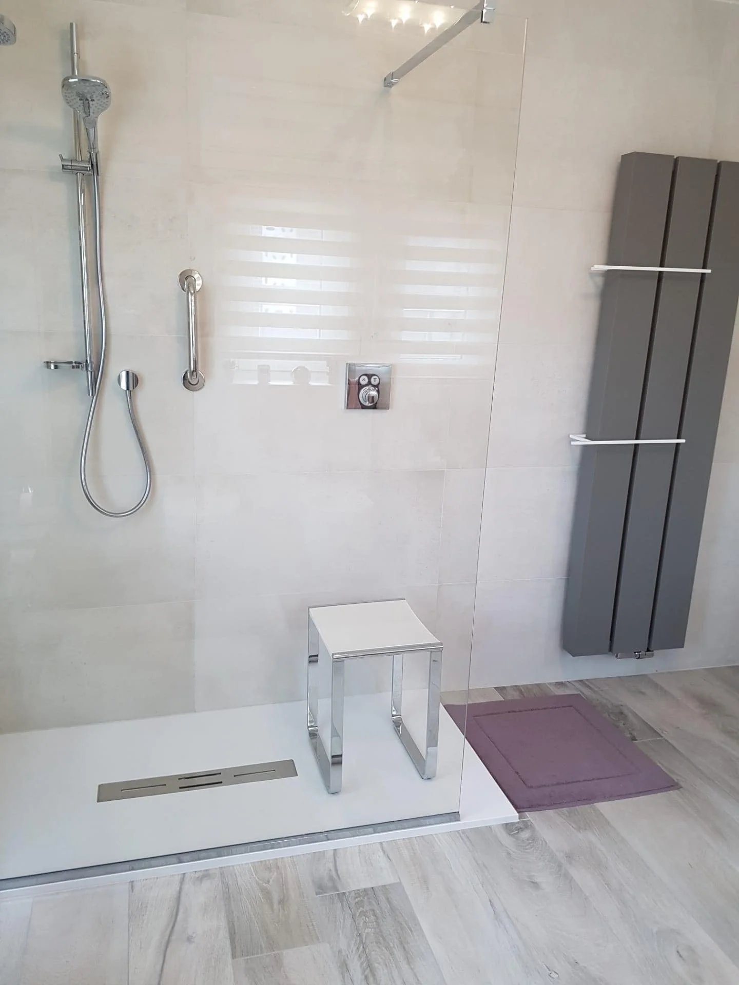 Een moderne, drempelloze inloopdouche met een glazen wand, een handdouche en een thermostatische mengkraan. Er staat een witte krukje en een kleine mat voor de douche, en een strakke, verticale kast aan de muur.
