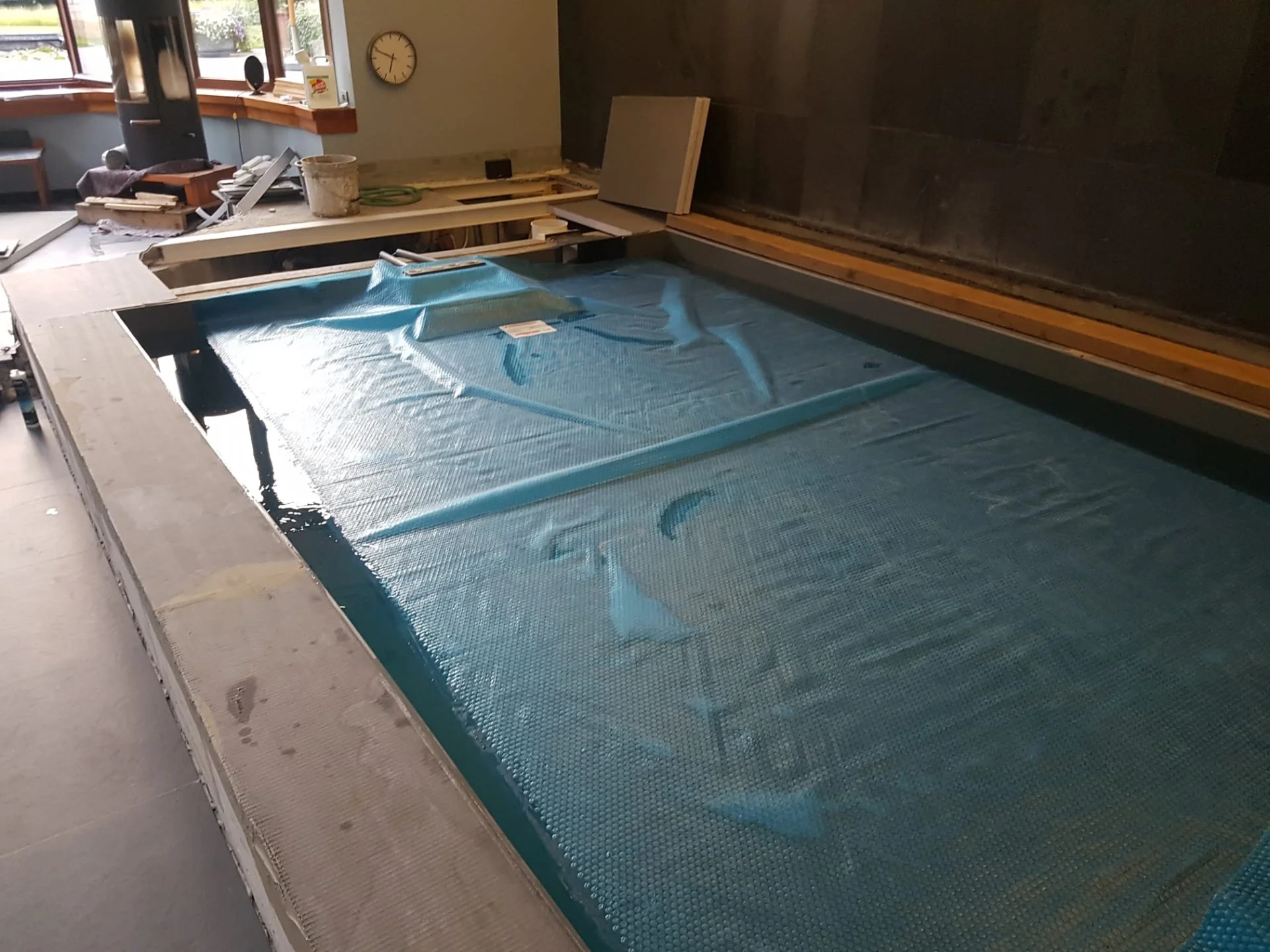 Binnenkant van een zwembad dat wordt aangelegd of gerepareerd, bedekt met een blauwe beschermlaag of net, in een kamer met een raam en een wandklok.