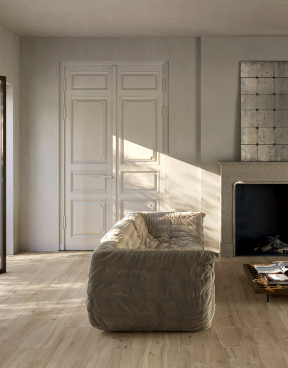 Een woonkamer met een loungebank, een open haard en een deur achter het raam die open lijkt te staan. Zonslicht valt op de muur en de bank.