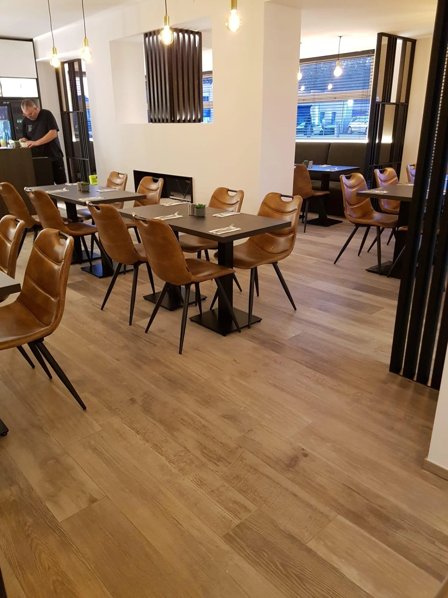 Een modern restaurantinterieur met houten vloeren, meerdere houten stoelen en zwarte tafels. Er hangen ingebouwde lichtarmaturen aan het plafond, en grote ramen laten het daglicht binnen. Er is een persoon die in de keuken bezig is.