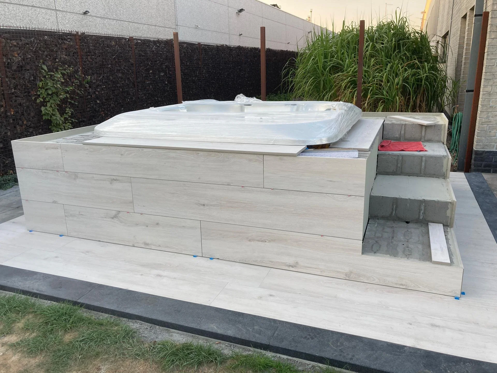 Een in aanbouw zijnde jacuzzi op een verhoogd terras, omgeven door betegelde veld en groene planten op de achtergrond.