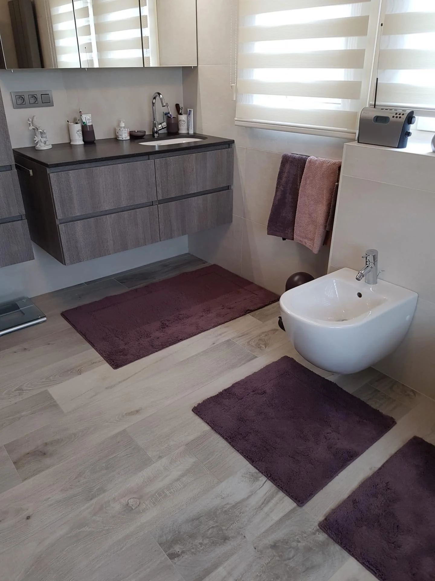 Een moderne badkamer met een muurhangende wastafel, paarse badmatten, een bidet, handdoeken, een spiegel en een raam met jaloezieën.