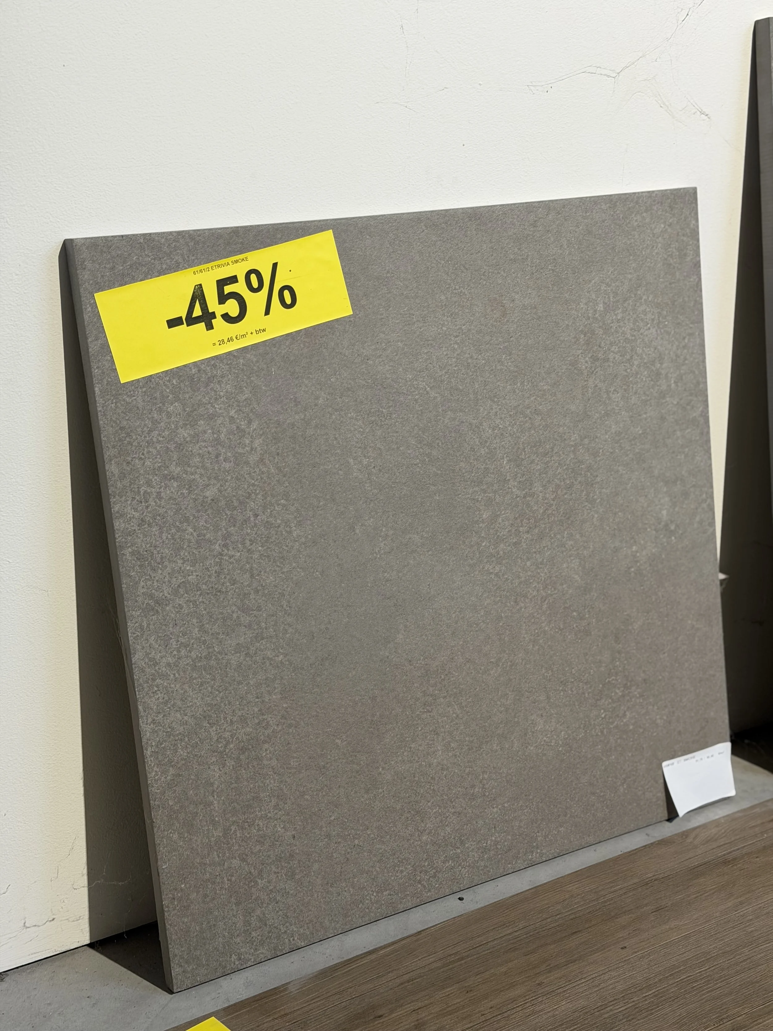 Een hoek van een bouwmuur met een grijstinten muurpaneel en een gele uitverkoopsticker met 45% korting. Op de onderkant ligt een houten vloer.