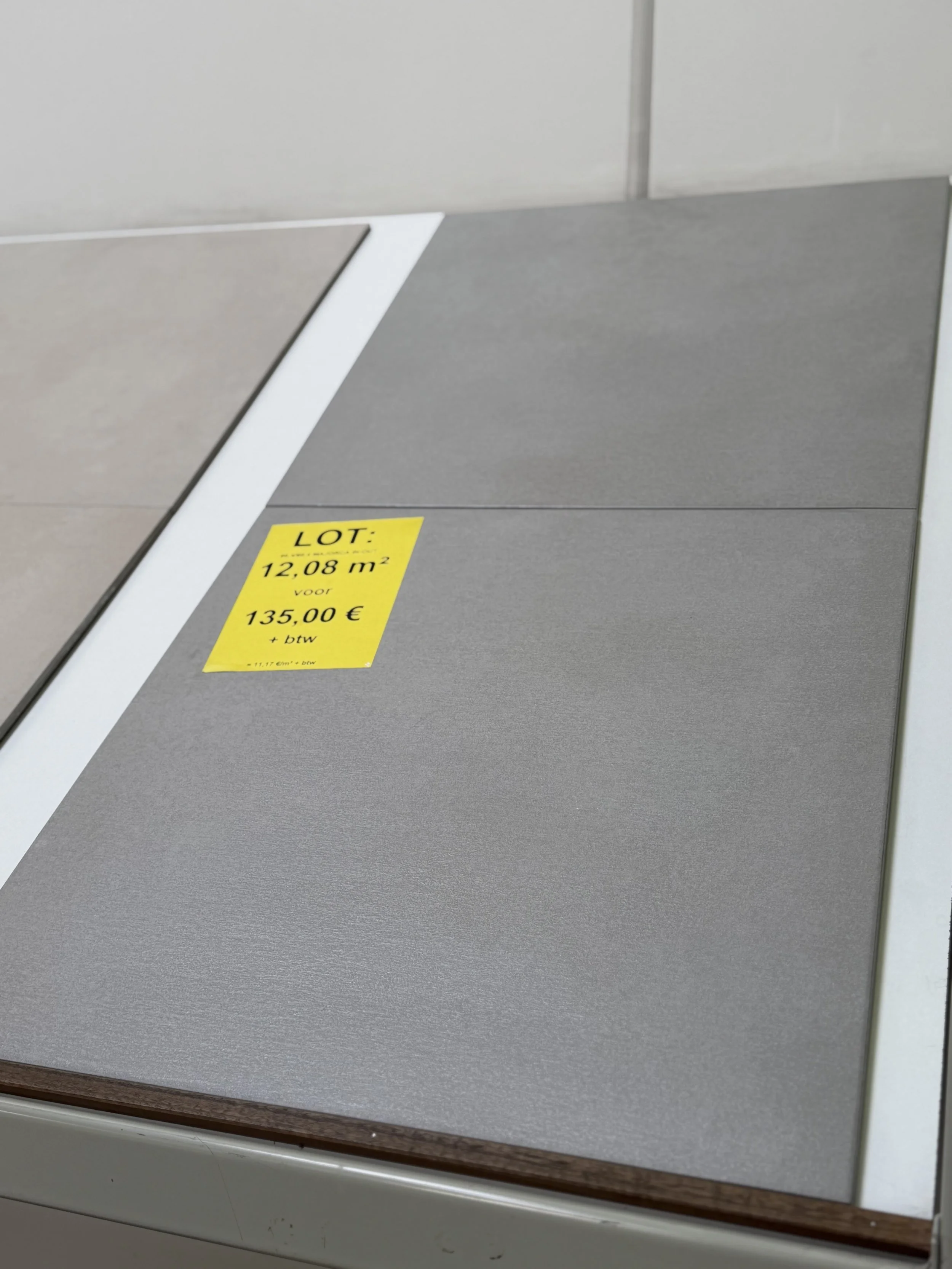 Verschillende tegels in grijstinten uitgelijnd op een vloer of muur, met een gele sticker die informatie bevat over het lot, oppervlakte en prijs.
