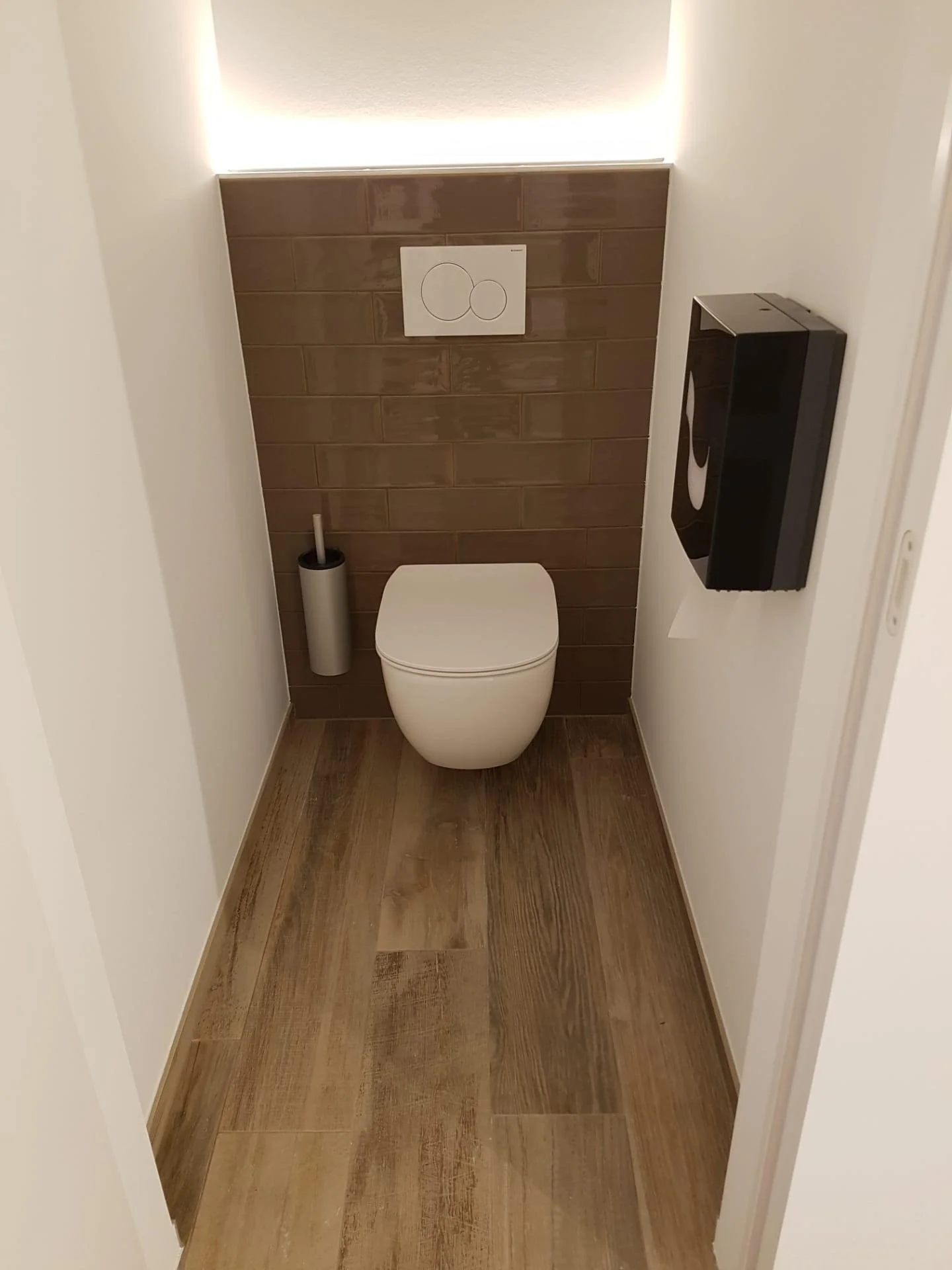 Moderne toiletruimte met witte toilet en houten vloer, een houten wand met een inbouw-wc-flush bediening en een toiletborstel, afgewerkt met een ingebouwd licht boven de muur.