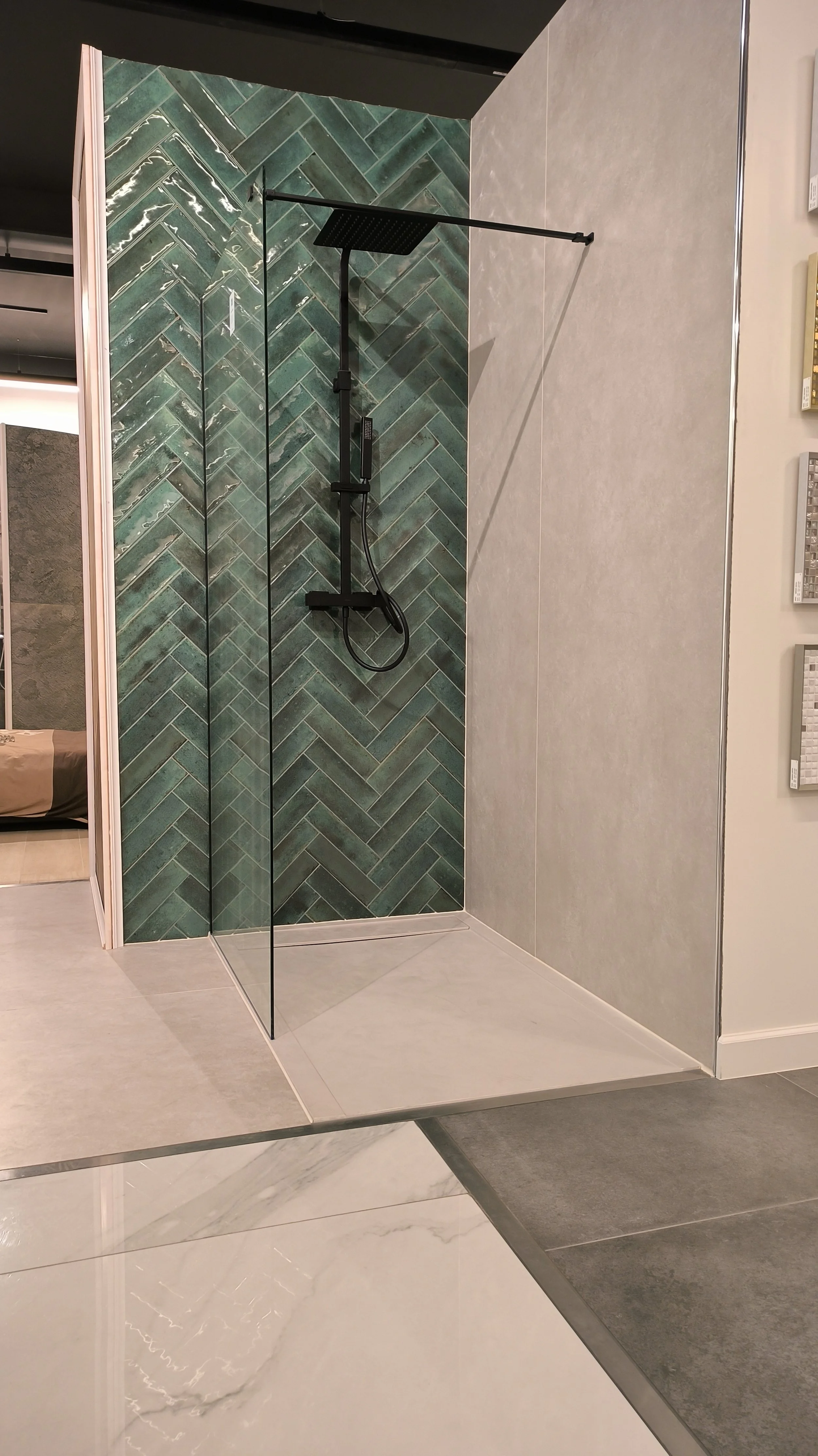 Een douchecabine met zwarte sanitaire voorzieningen, waaronder een hoofddouche, in een badkamer met groene tegelmuur in een visgraatpatroon. De vloer buiten de douche is lichtkleurig en de muur buiten de douche is lichtgrijs.