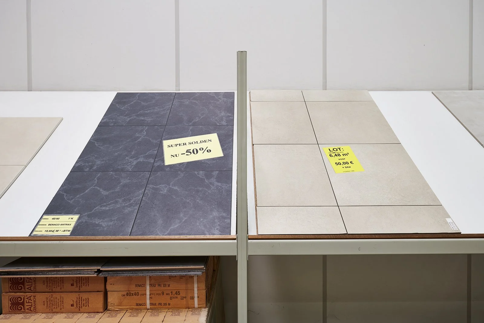 Tegels in een winkel, links donkergrijs met marmerpatroon, rechts beige, beide op een witte tafel met prijs- en promotieborden.
