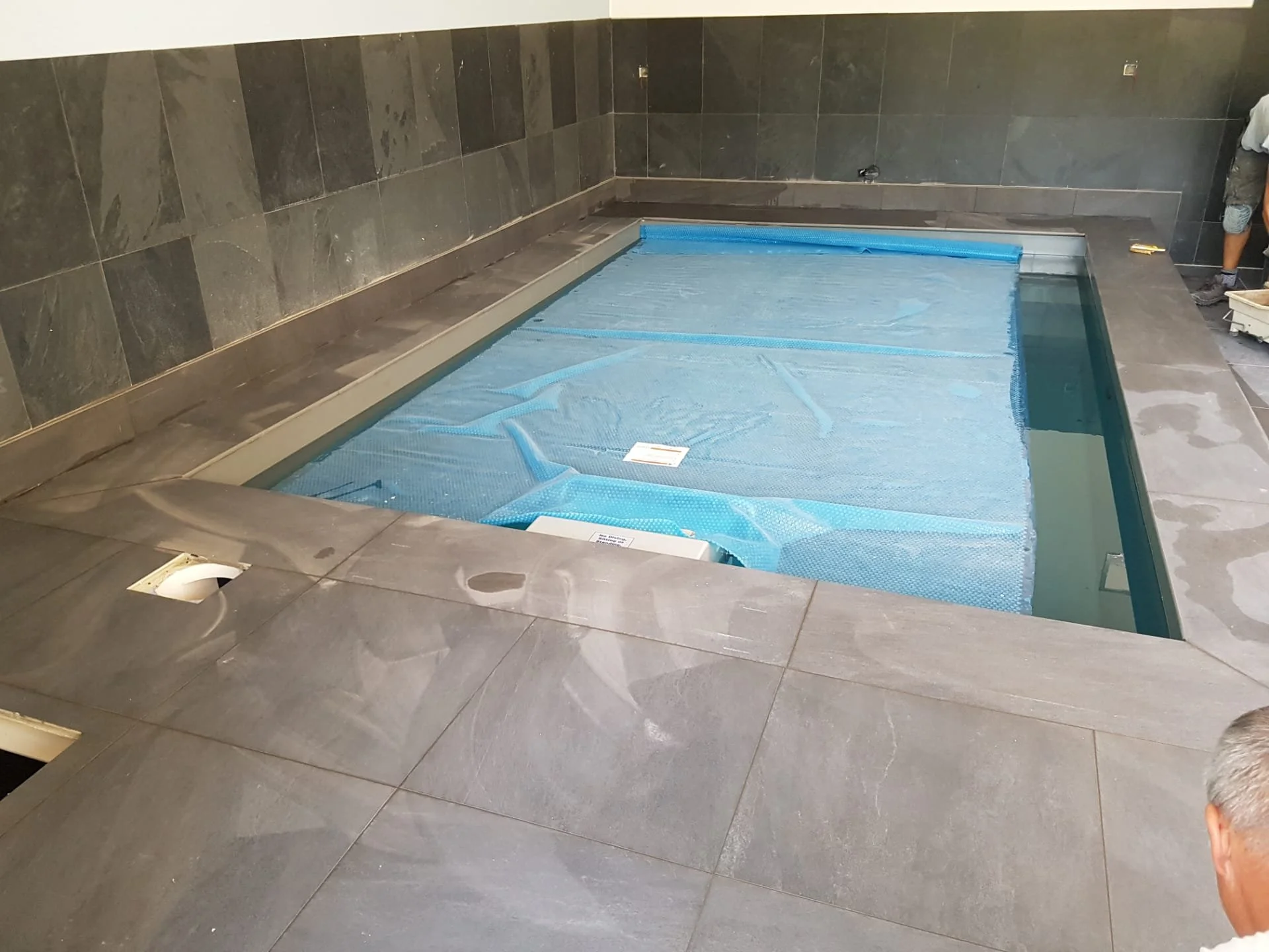 Een zwembad in aanbouw met water dat wordt getest, omgeven door een grijze tegelvloer en donkere muurranden. Een persoon staat aan de rechterkant, gedeeltelijk zichtbaar.
