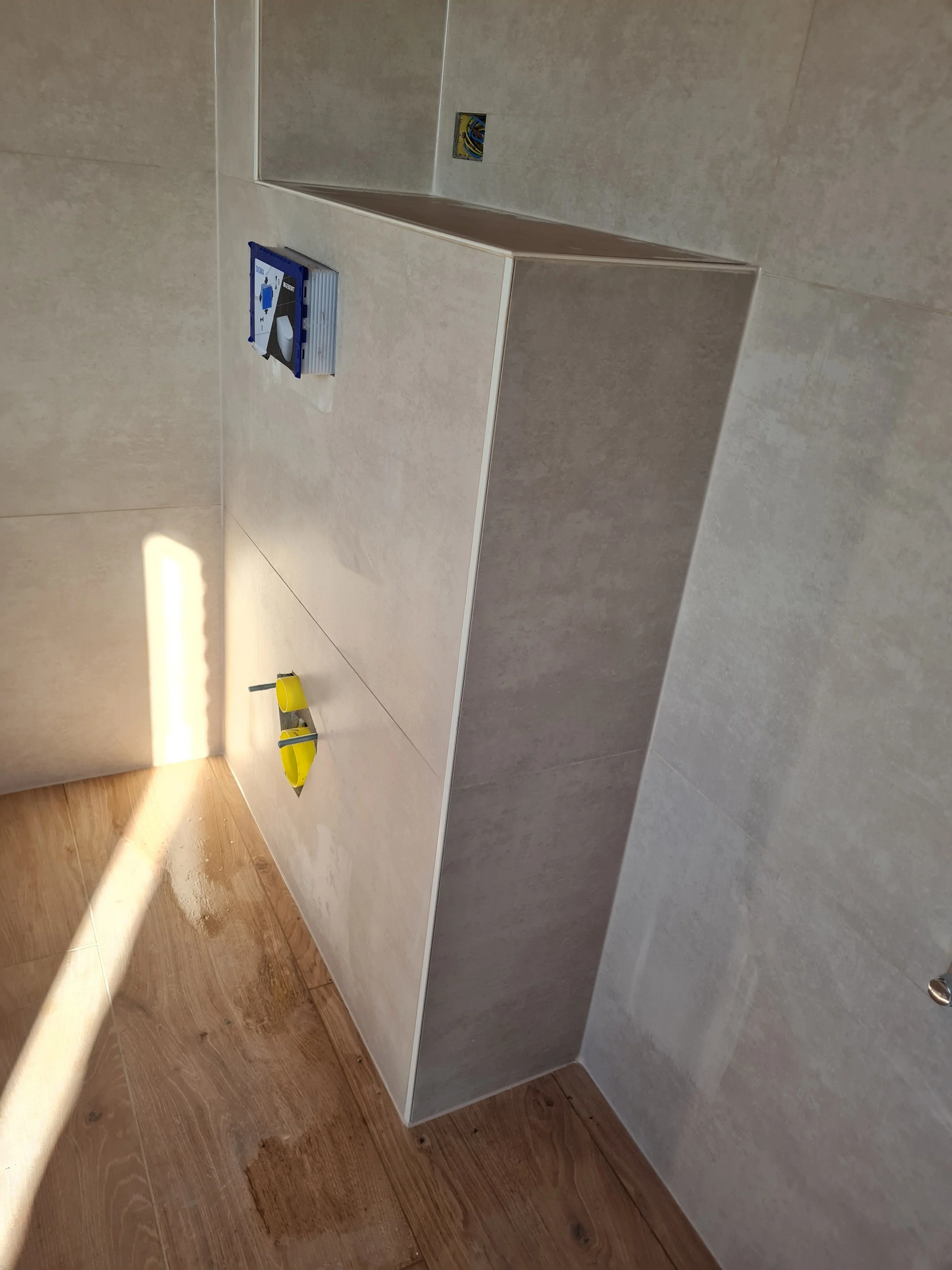 De afbeelding toont een open lade in een keuken of badkamer, met een elektrische aansluitdoos en een stuk gereedschap. Er ligt ook een gele emmer en een ijzeren haakje op de vloer.