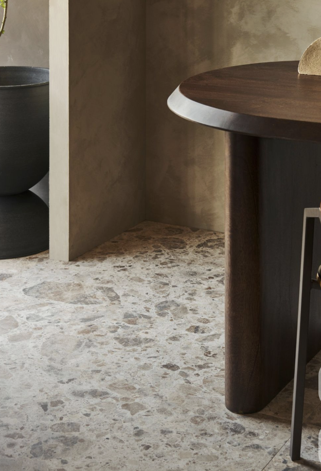Een hoek van een kamer met een terrazobodem, een donkere ronde tafel met een houten blad, een zwarte pot en een muur met een natuurkleurige afwerking.