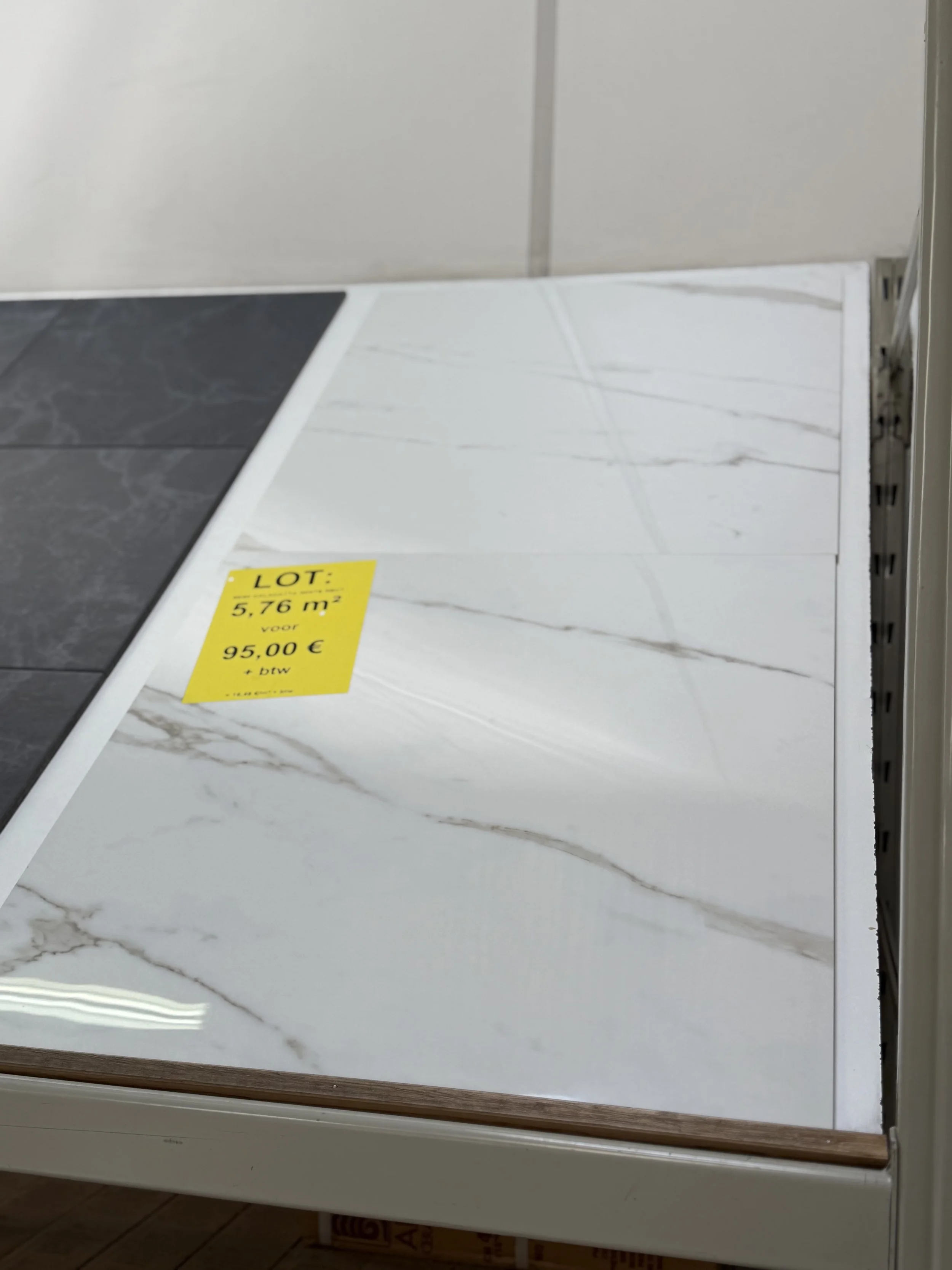 Twee kleuren tegels op de vloer, een donkere en een lichte met een witte marmerachtig patroon, met een gele prijskaartje dat 5,76 m² en €95,00 vermeldt.