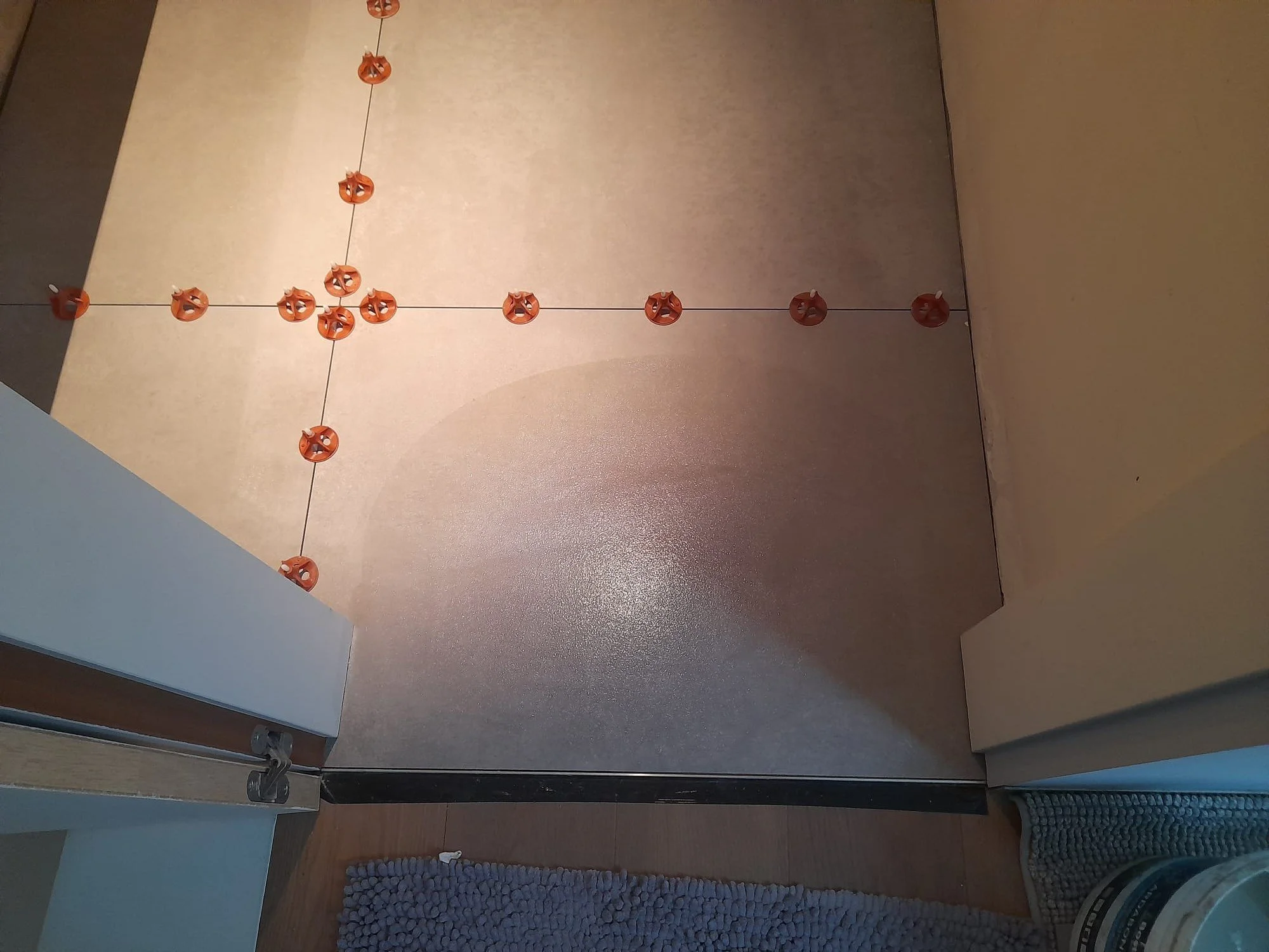 De afbeelding toont een hoek van een keuken met een tafel of keukeneiland. Op het aanrecht liggen oranje kurkentrekkers, die in een patroon liggen. Op de vloer ligt een witte badmat.