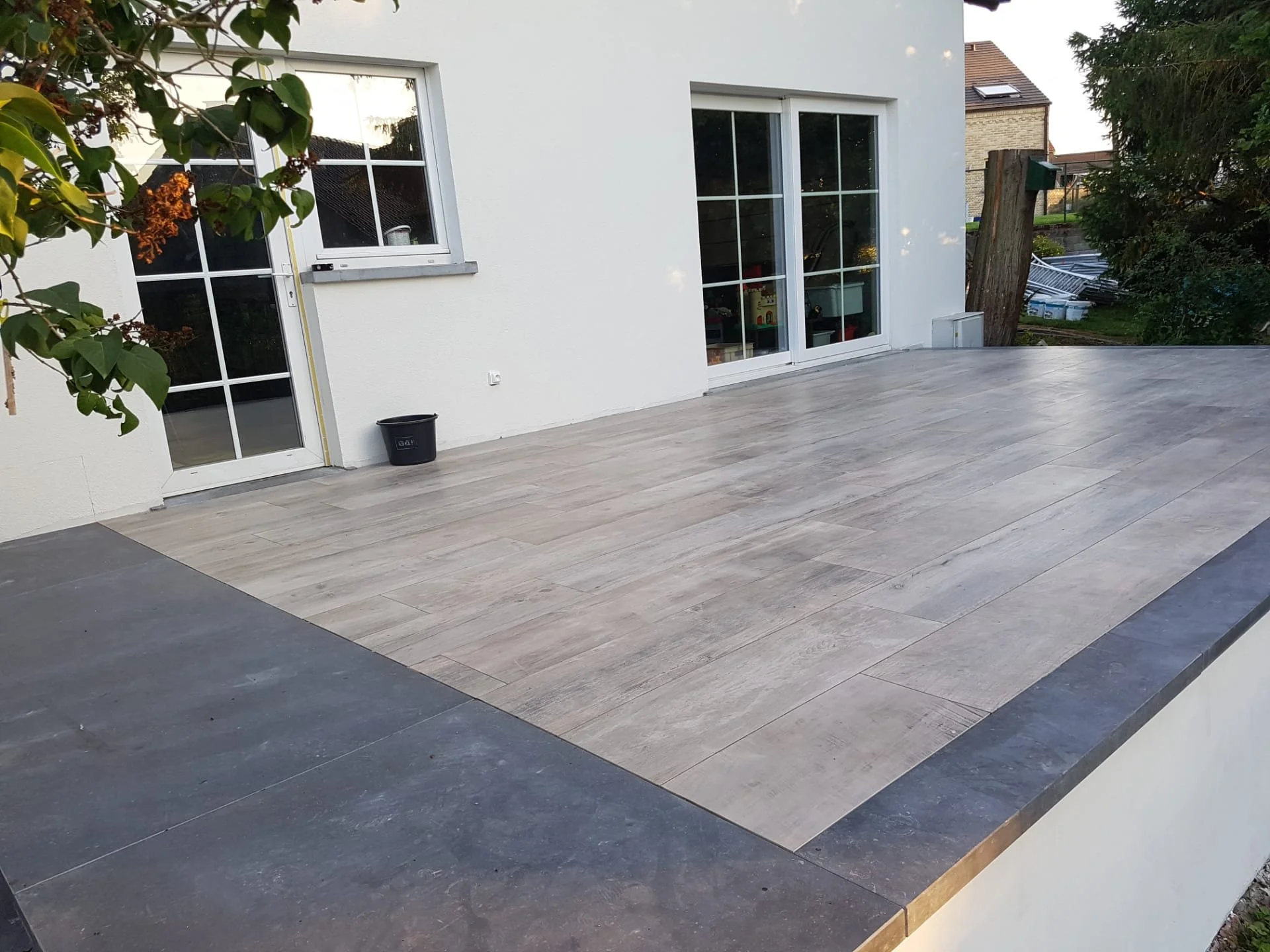 Een nieuw aangelegde terras met grote donkergrijze en lichtgrijze tegels, aan de muur van een wit huis met drie grote ramen en een deur met raam.