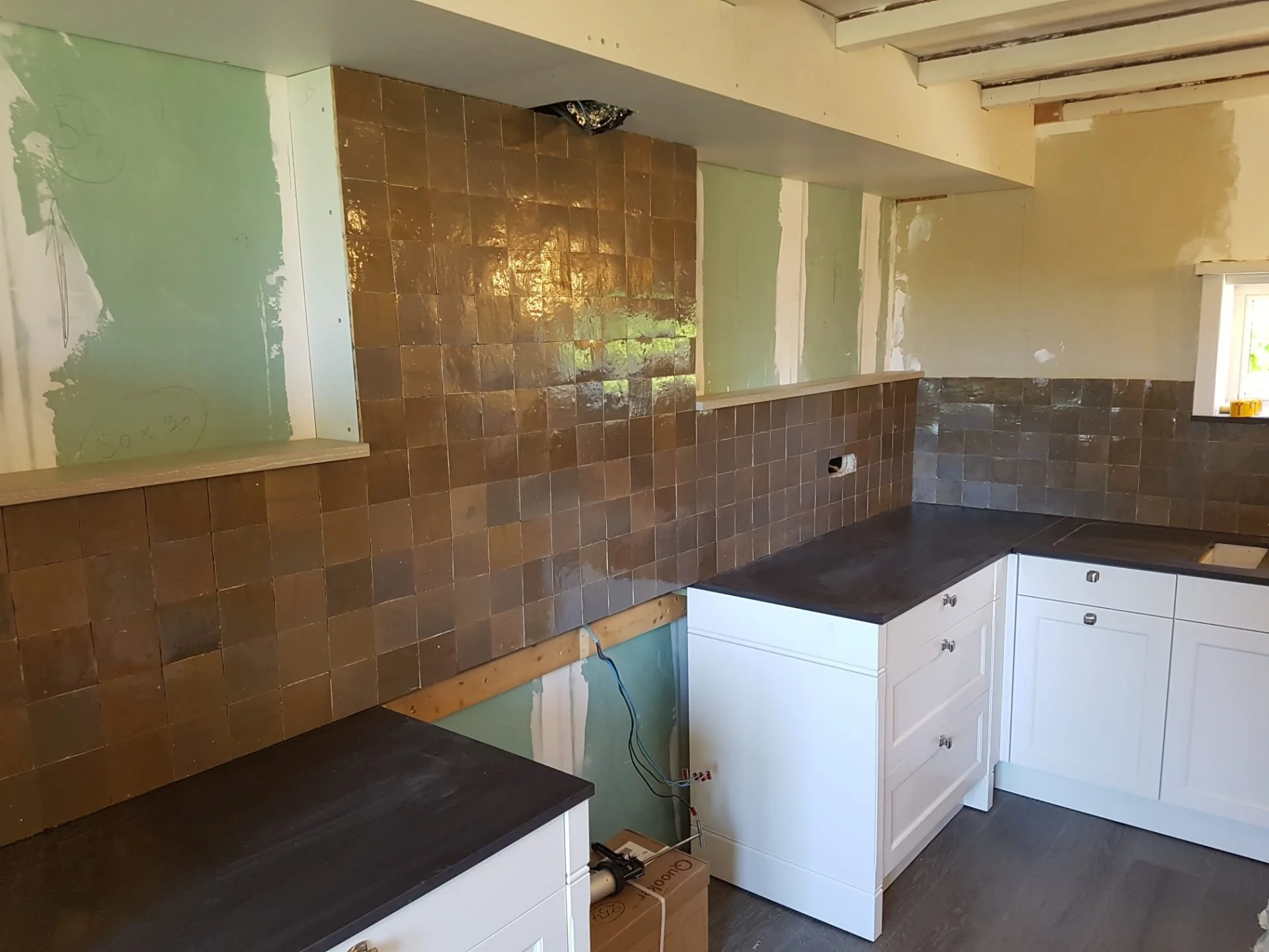 Een keuken onder renovatie met bruin gebakken keramische tegels op de muur en zwarte werkbladen. Er is een wit kastje onder een zwart werkblad en een kist op de vloer. Een raam laat natuurlijk licht binnen.