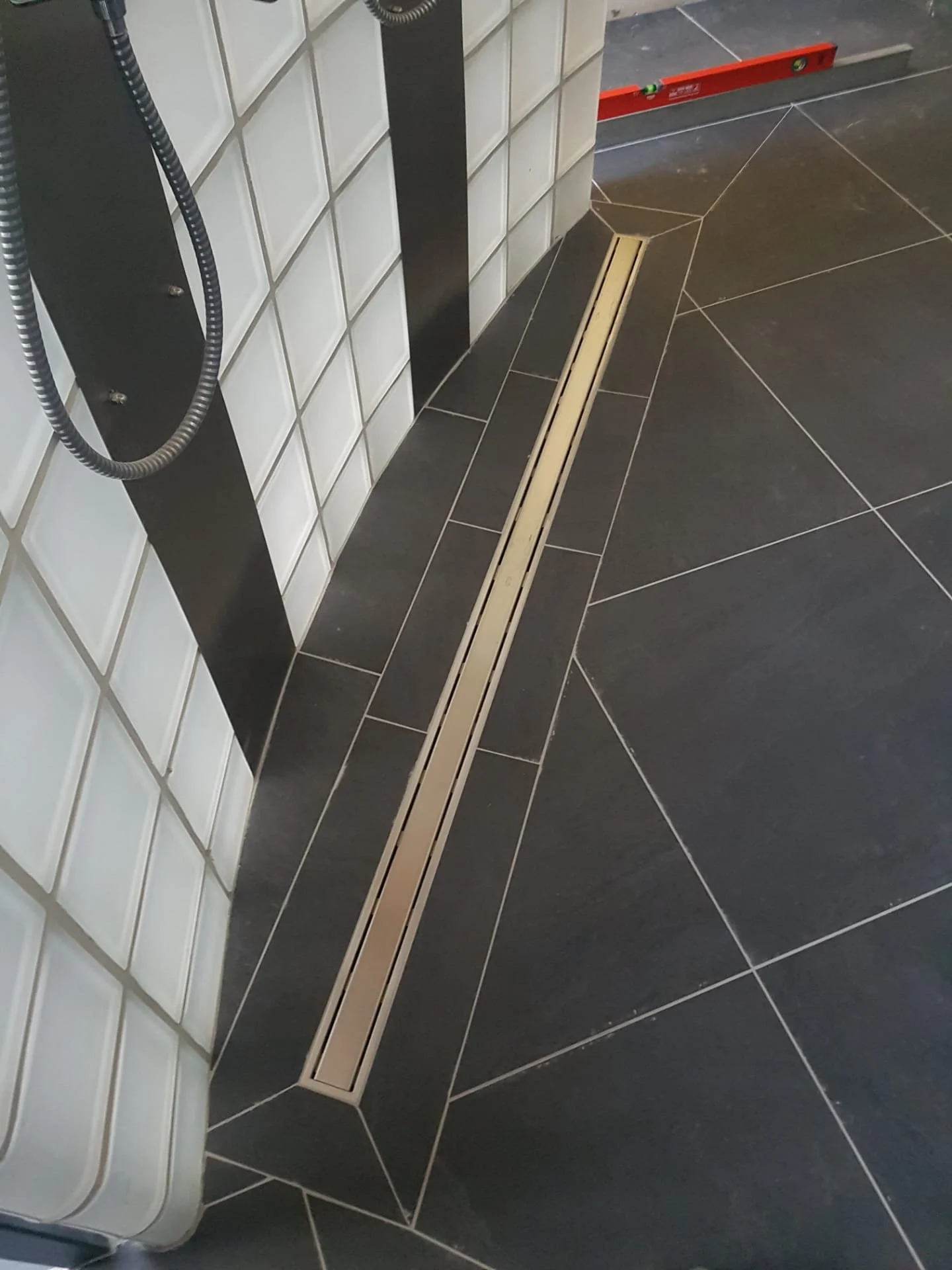 De afbeelding toont een vloer met zwarte tegels en een lange, smalle afvoergoot. Linksboven bevinden zich enkele groene en rode instrumenten, en een zwarte muur met witte bakstenen. Links hangen zwarte en grijze snoeren of kabels.