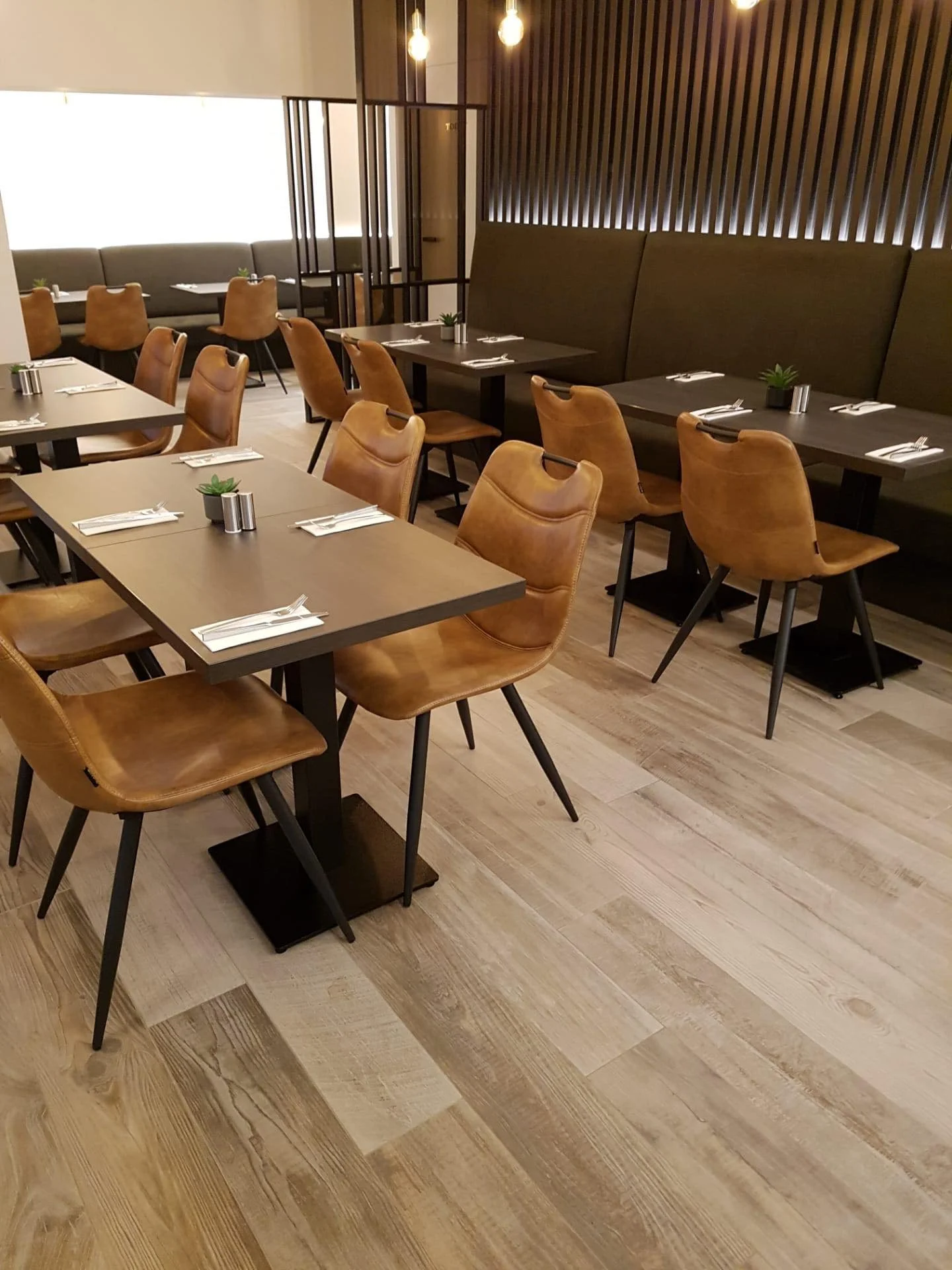 Een restaurantzaal met houten vloeren, meerdere tafelinstellingen en leren stoelen, kleine planten op de tafels en warme verlichting.
