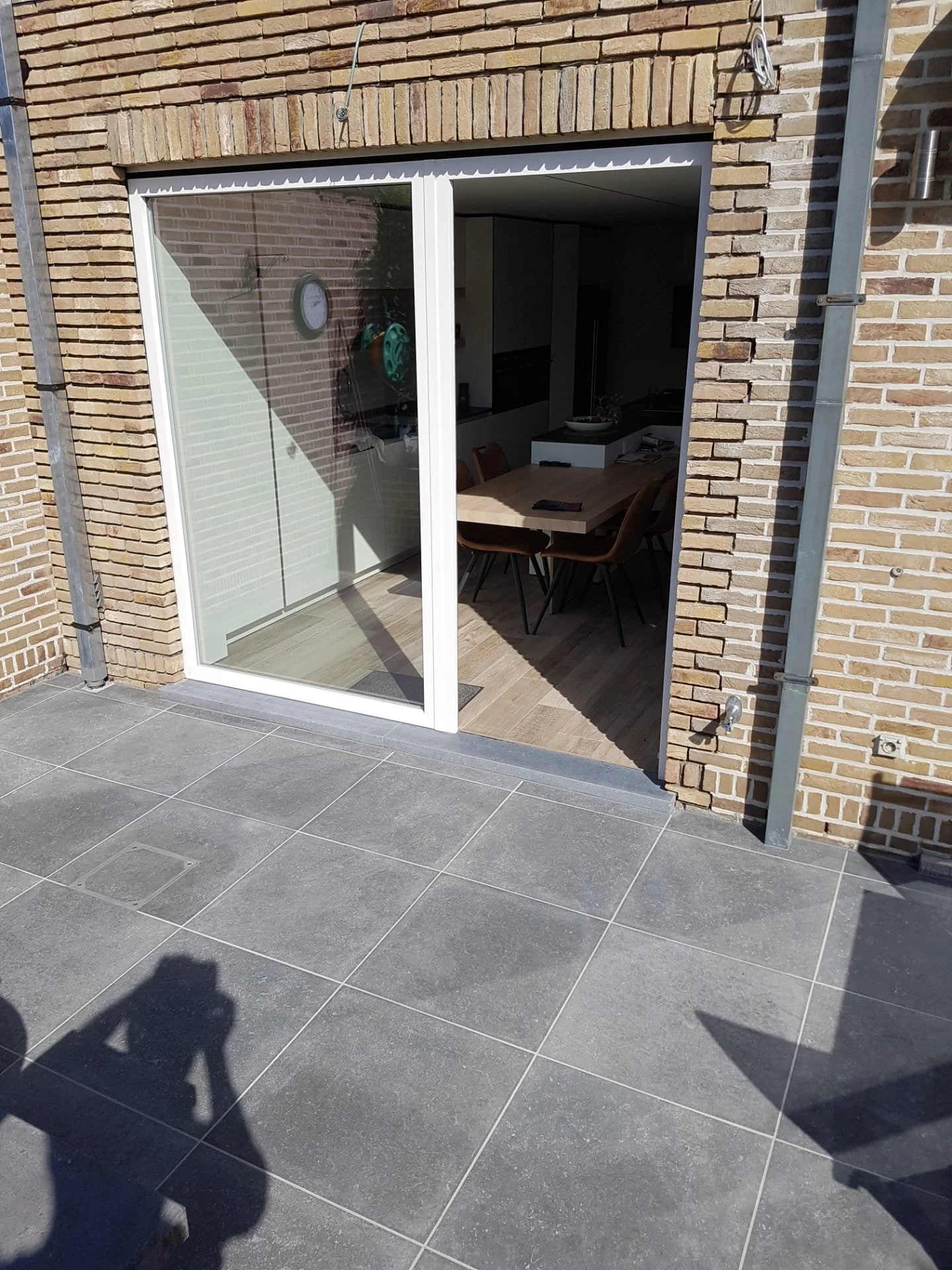 Gevel met grote glaspui, binnen een tuin met grijze tegelvloer en een muur van bakstenen.