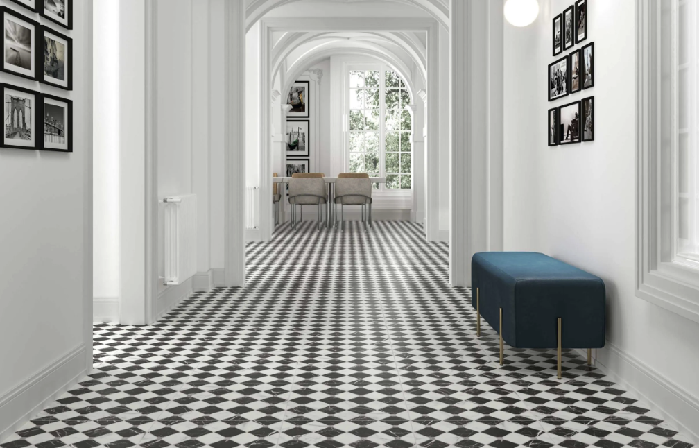 Een wit geschilderde gang met zwart-wit geometrische tegelvloer, foto’s aan de muren, een blauwe bank aan de rechterwand en een deur die naar een andere kamer leidt met een grote raam en een eettafel met drie stoelen.