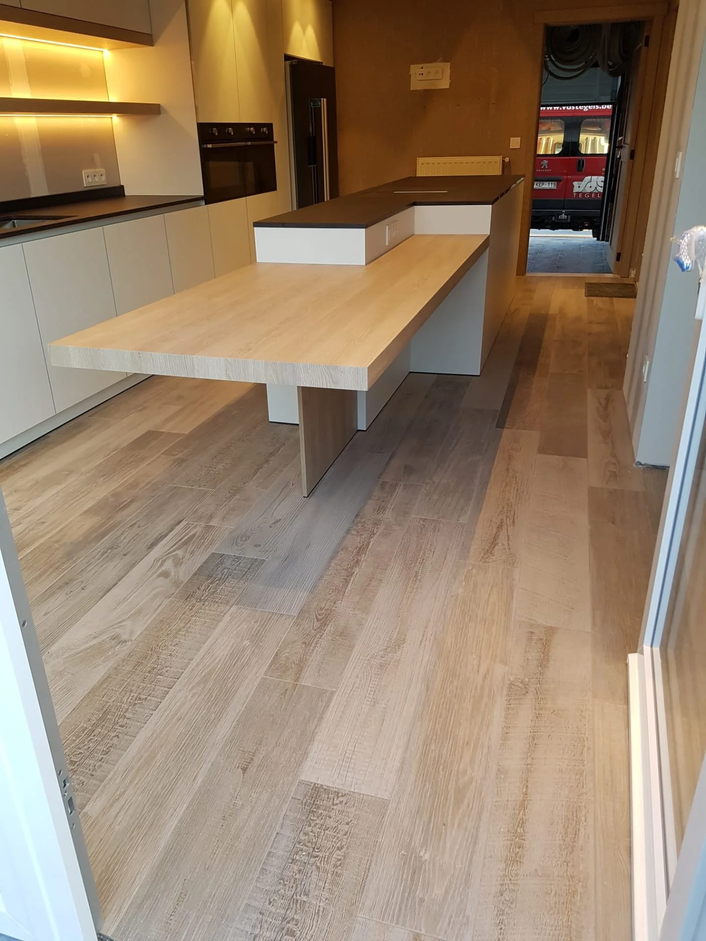 Een moderne keuken met een houten eettafel en beige keukenkasten, een oven, een koelkast, en een deur die uitkomt op een straat met een vrachtwagen.