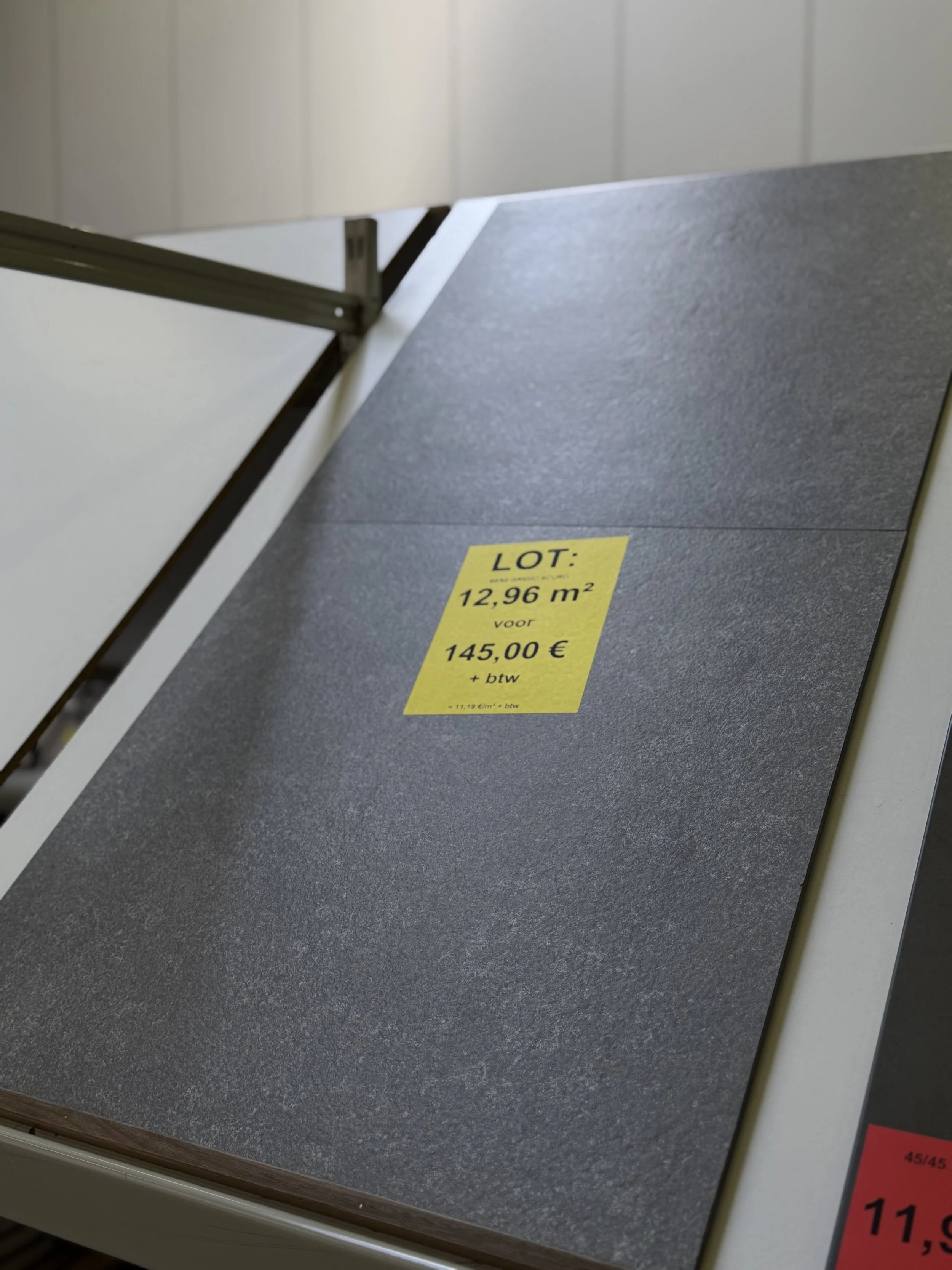 Een zwarte markeer met een gele sticker die informatie bevat over een kavel van 12,96 vierkante meter voor 145 euro. De foto toont een vloer met een rand en een deel van een muur.
