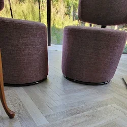 Twee ronde, gestoffeerde stoelen in een modern interieur met houten vloer, naast een grote raam met uitzicht op groen outdoor.