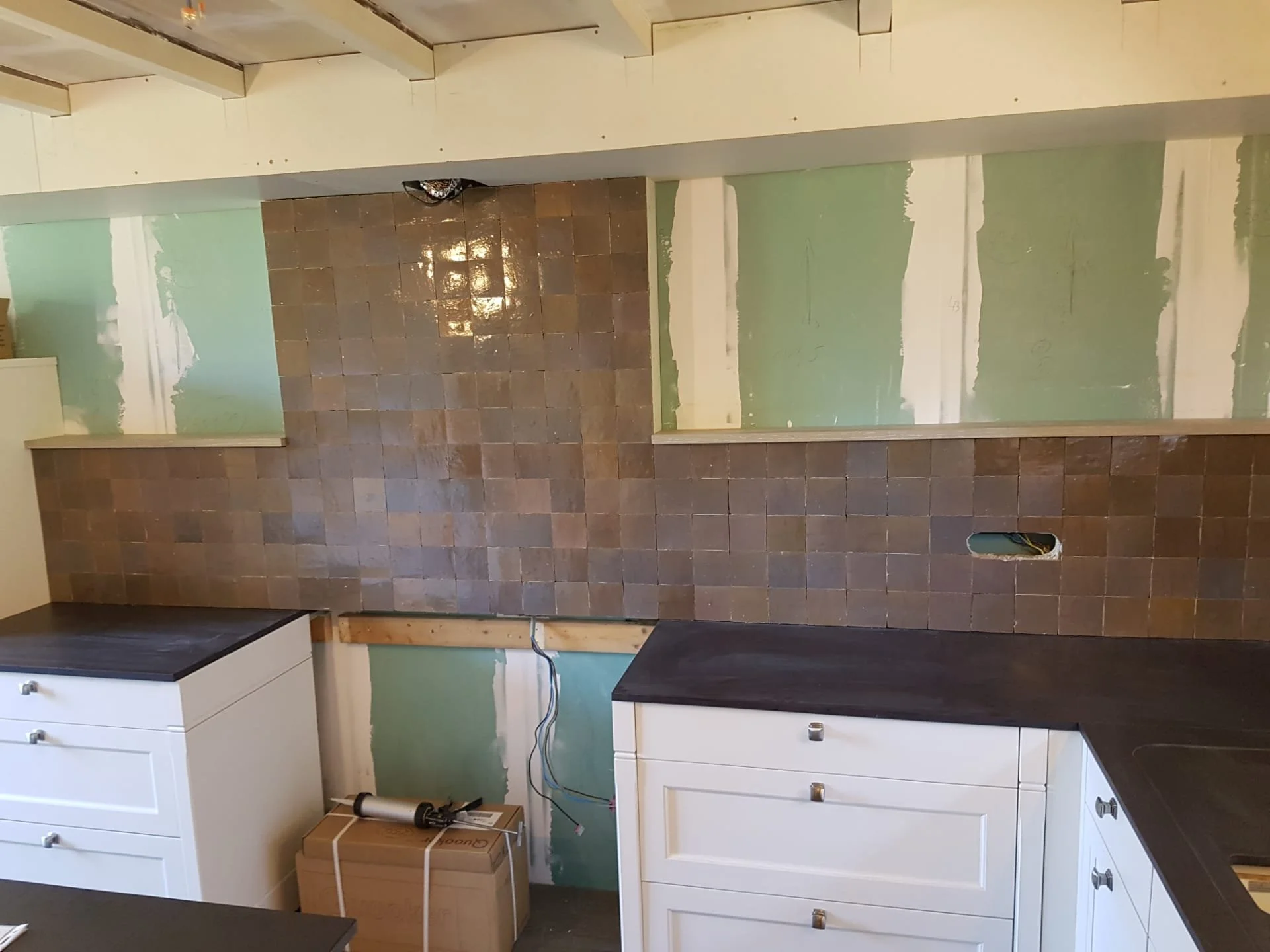 Deze afbeelding toont een keuken in voorbereiding voor renovatie, met een nieuwe bakkolom van bakstenen en een muur met groene loodverf en droge bekisting. Er staan wit gelakte kasten onder de muur, en er ligt een doos met bouwmaterialen op de vloer.