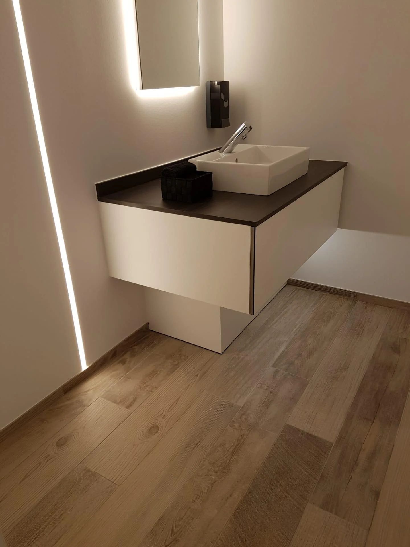 Een wit wasbakje op een zwart blad met een handdoekje en dispenser ernaast, in een modern badkamer met houten vloer.