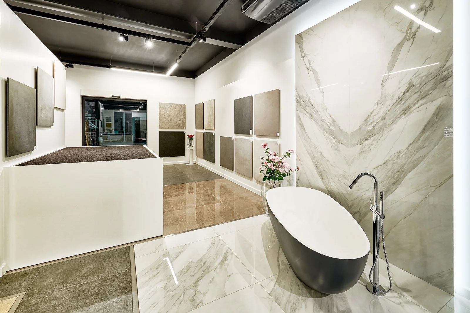 Een moderne, minimalistische badkamer met een zwarte vrijstaande badkuip, omringd door marmeren wanden en vloeren, en een collectie van neutrale gekleurde wandpanelen of tapijten aan de muur. Decoratieve bloemen en inbouwverlichting voegen een luxe u