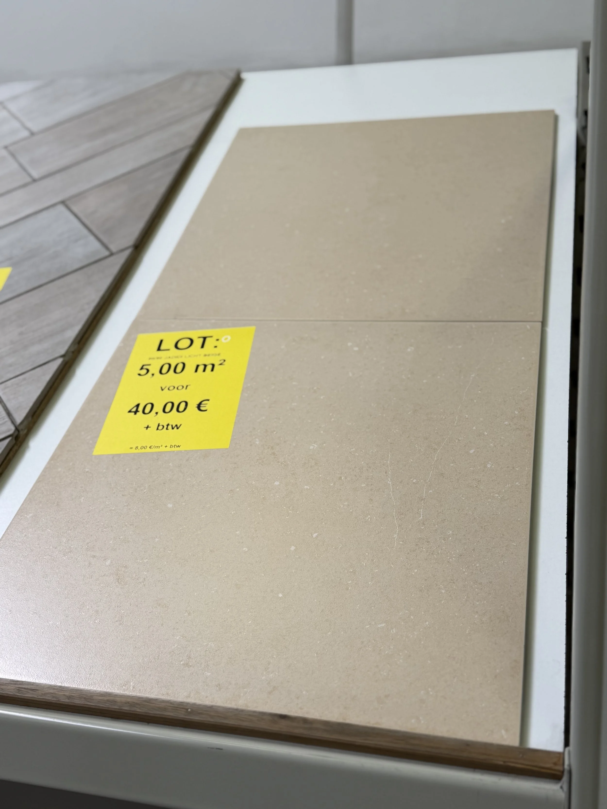 Close-up van een stuk beige vloer- of wandtegel met een gele koopwaar-prijskaart erop, waarop staat: LOT: 5,00 m² voor 40,00 € + btw.