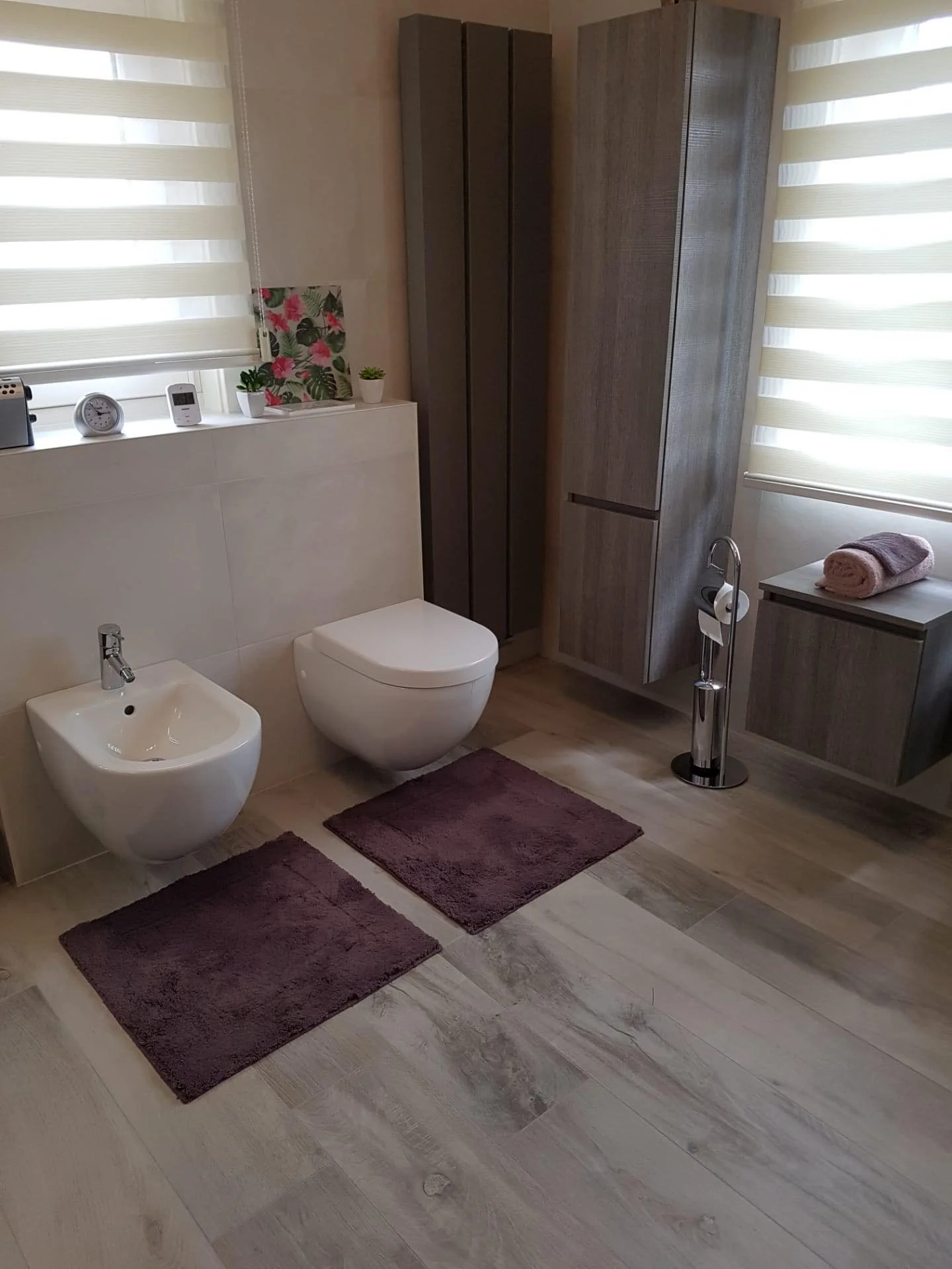 Een moderne badkamer met wandcloset en bidet, twee paar paarse vloerkleden, een houten kast en een kast met handdoeken, twee ramen met horizontale jaloezieën, decoratieve planten en een klok op het vensterbank.